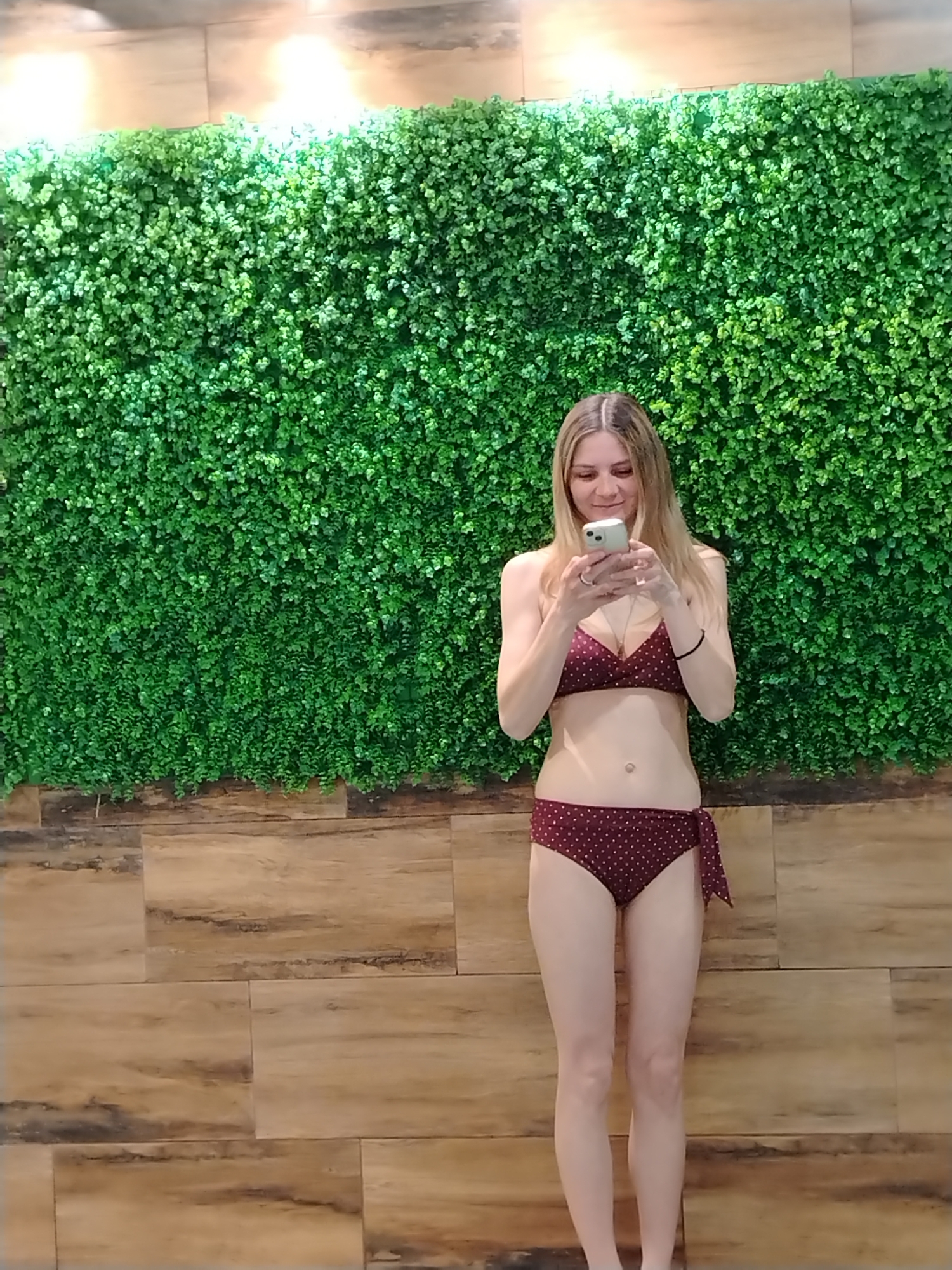 A woman stands in front of a wall composed of artificial green plants above wood-like tiles. She is wearing a two-piece bikini with a burgundy and gold polka dot pattern and is holding a smartphone, looking at the screen. The photograph was taken indoors in Chaikovsky, Russia.