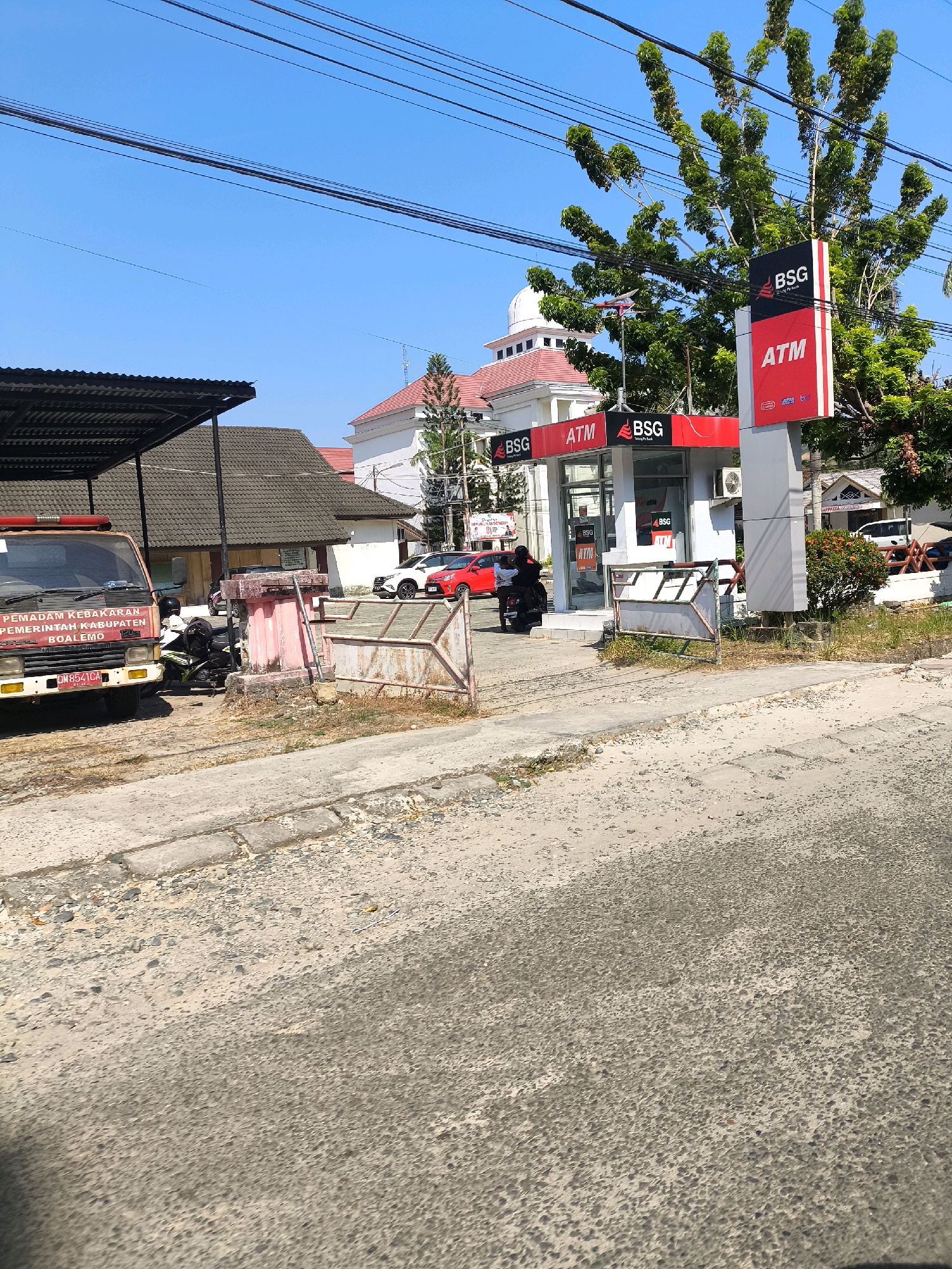 The image depicts an outdoor scene in Tilamuta, Indonesia, featuring a BSG Bank ATM booth and surrounding area. In the foreground, a paved road with a concrete curb is visible, showing signs of wear and tear with loose gravel. To the left, a red and white fire truck with the text "PEMADAM KEBAKARAN PEMERINTAH KABUPATEN BOALEMO" is parked under a corrugated metal awning. Adjacent to the fire truck, a motorcycle with a rider is parked.
The central focus is a modern BSG Bank ATM booth with red and black signage. Two ATM machines are visible within the glass-fronted structure. A tall advertising stand for BSG ATM is positioned to the right of the booth. Behind the ATM booth, a white building with a dome and red roof is partially visible, possibly a government building or place of worship. Several trees with green foliage are present, with one prominent tree behind the ATM stand. In the background, other buildings and a few parked cars, including a white SUV and a red car, are discernible. The sky is clear and blue. The overall environment suggests a sunny day in a semi-urban or commercial area.
