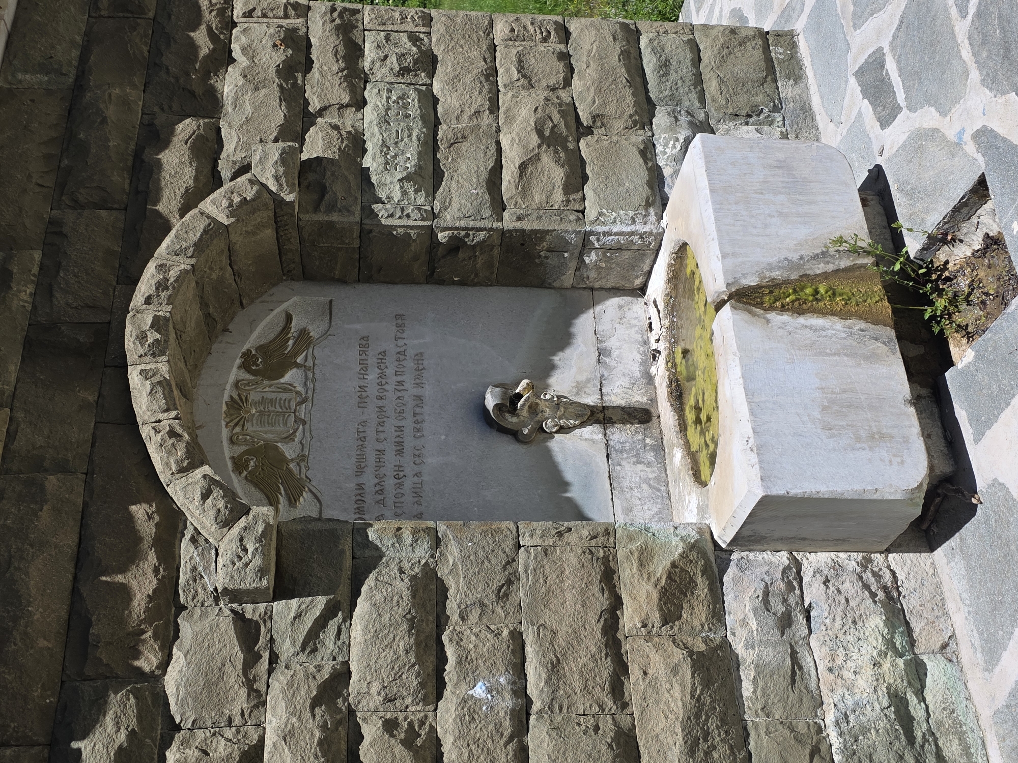 The image shows a stone fountain in Bachkovo, Bulgaria. The fountain is set into a stone wall, with a rounded archway above it. There is an inscription on the wall above the fountain's spout. The fountain itself is made of white stone and has a pool of water in the basin. Plants are growing around the fountain. The surrounding area is paved with stone.
