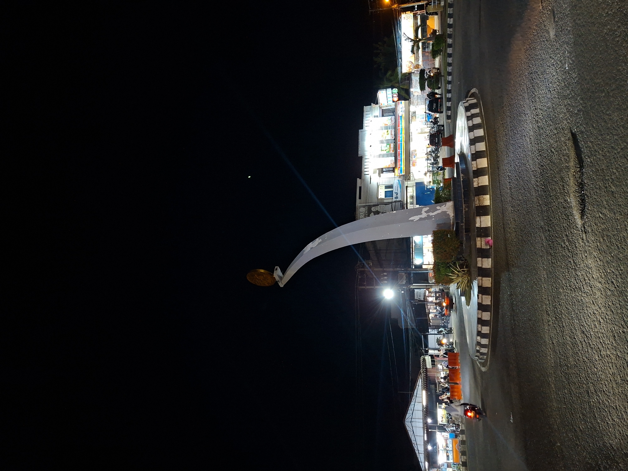 The image depicts a street scene in Lhokseumawe, Indonesia, at night. A major thoroughfare is bordered by illuminated shops and restaurants. A large, white archway with a circular ornament stands prominently on the side of the road. Cars and motorcycles are seen travelling on the road. The night sky is dark and clear, with a small, faint celestial object visible.
