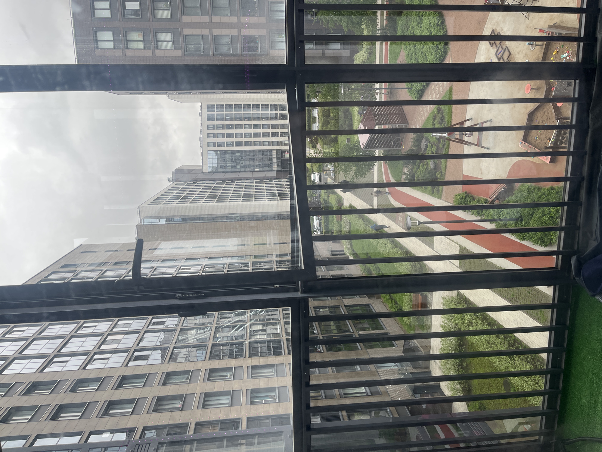 The image is taken from a high vantage point, likely from inside an apartment building in Saint Petersburg, Russia, looking out through a window with a metal railing. The view is angled downward towards a courtyard or recreational area. In the foreground, the metal railing of the window is visible. Beyond the railing, the courtyard features a playground with a sandbox and climbing structures, interspersed with green landscaping, including bushes and trees. There are pathways made of concrete and red pavement. Several apartment buildings with numerous windows surround the courtyard. The sky appears overcast. No people are clearly visible in the courtyard, but there may be some figures in the distance. There is a patch of artificial grass on the right side of the image, inside the balcony.