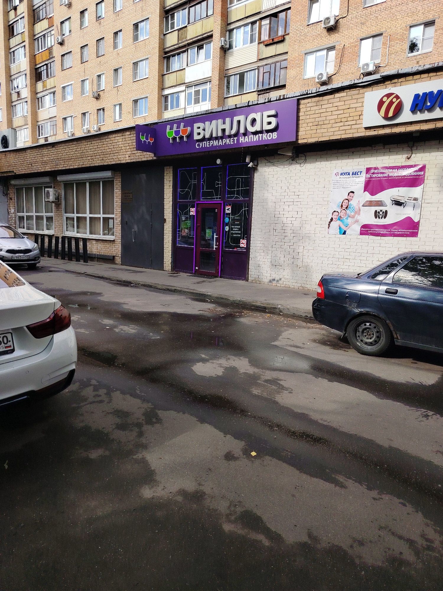 The image shows a street scene in Dolgoprudny, Russia. In the foreground, a white car is parked on the left, partially visible from the rear. To the right of the white car, a dark-colored sedan is parked. The street is wet, indicating recent rain, with puddles reflecting the sky and surrounding buildings.
In the background, a brick building houses commercial establishments. A prominent purple sign with white lettering reads "ВИНЛАБ" (VINLAB) and "СУПЕРМАРКЕТ НАПИТКОВ" (SUPERMARKET OF DRINKS). Next to the VINLAB sign, another sign with a red and white logo and blue lettering reads "НУГА БЕСТ" (NUGA BEST). A promotional poster is attached to the white brick wall of the building, featuring images of a family and what appears to be a massage bed or similar product. The building has several windows and air conditioning units visible on its facade. Further in the background, another multi-story residential building is visible, with many windows and balconies. The overall impression is of an urban environment, likely a commercial or mixed-use area.