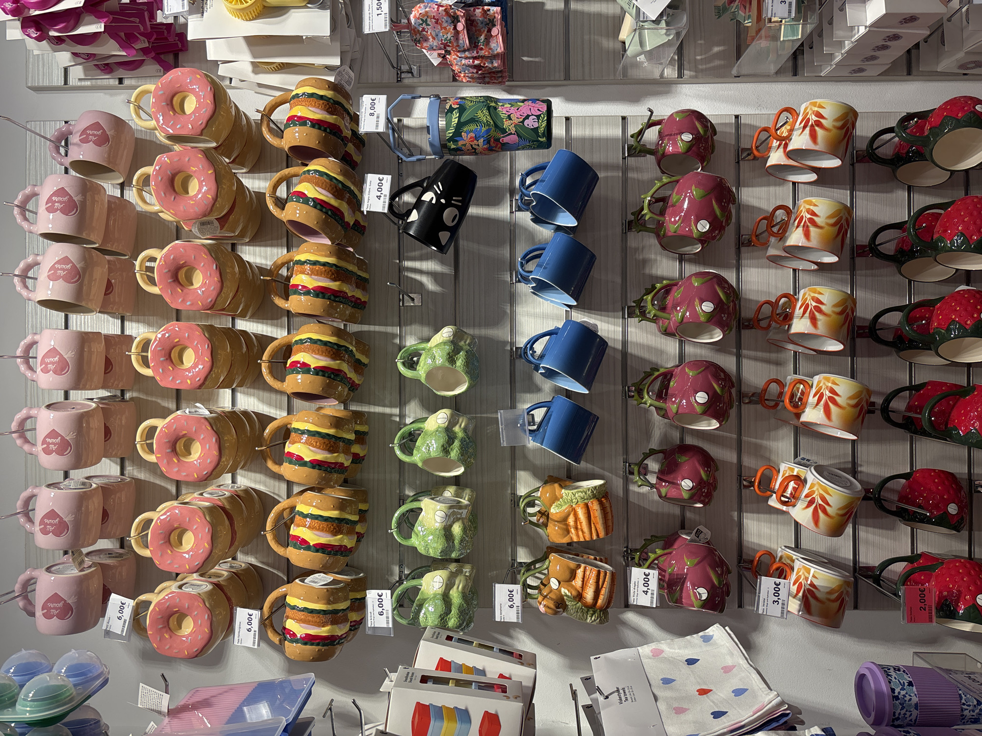 The image shows a wall display of novelty mugs in a store. The mugs are arranged in rows and feature various designs, including donuts, hamburgers, frogs, carrots, dragon fruit, strawberries, and a black cat. Some mugs have price tags, with prices ranging from €4.00 to €8.00. There are also some patterned fabric items and other merchandise displayed. The scene is brightly lit, suggesting an indoor retail environment. The specific location of Orihuela, Spain is not visible in the image itself.