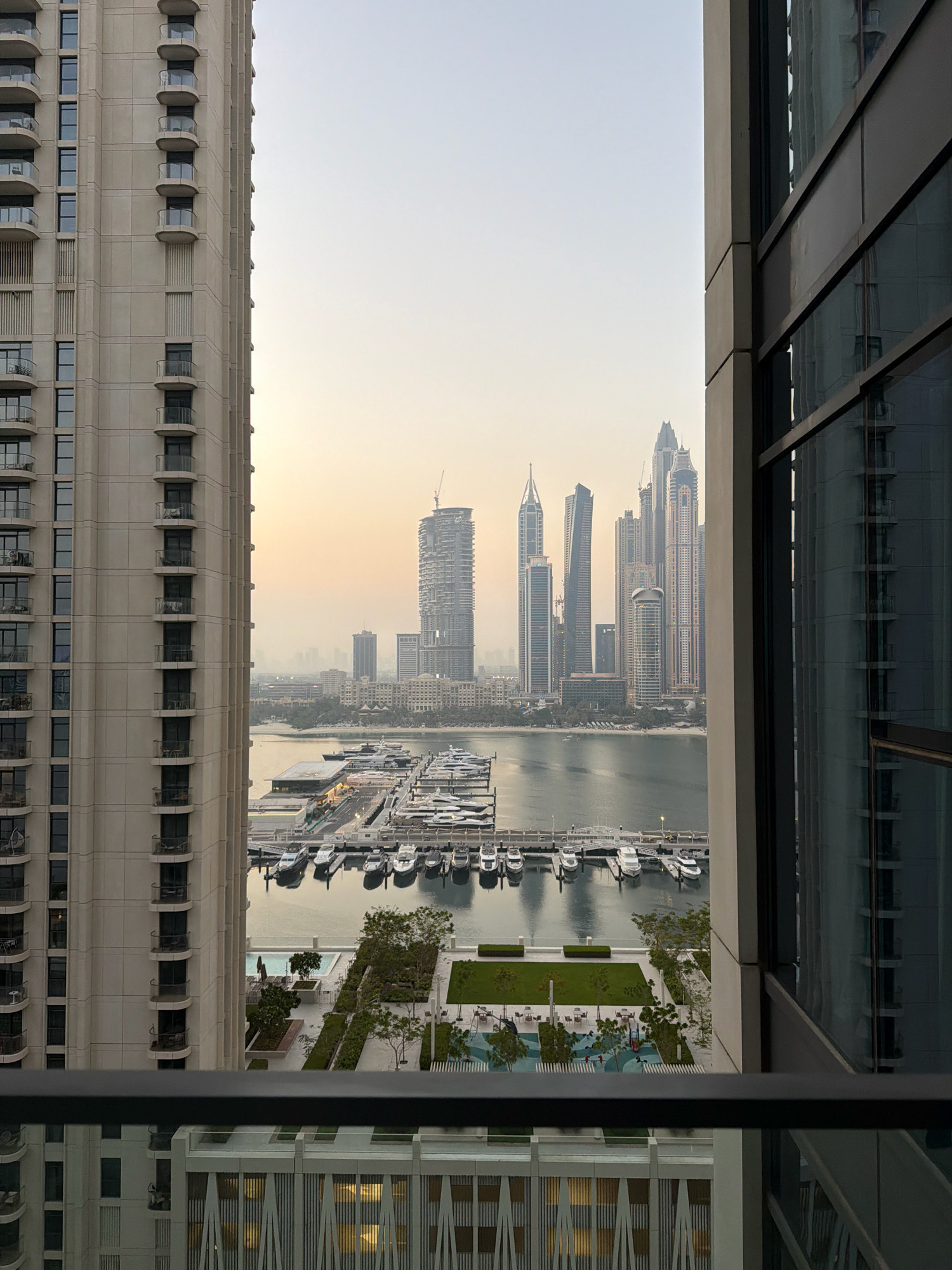 The photograph shows a view of the Dubai Marina in Dubai, United Arab Emirates. The foreground features a balcony with a railing, and the left side of the image is dominated by the facade of a tall residential building with many balconies and windows. To the right, another modern building with large, reflective windows is visible.
Beyond the buildings, the Dubai Marina is filled with numerous yachts docked in neat rows. A walkway or pier runs along the docks. In the middle ground, there is a landscaped area with green lawns, trees, a swimming pool, and a children's playground with colorful equipment.
The background showcases the iconic Dubai skyline, characterized by a dense cluster of very tall skyscrapers of varying architectural designs. The sky is a hazy, pale orange, suggesting either sunrise or sunset. The overall impression is an urban, contemporary scene with a focus on luxury waterfront living and modern architecture. No people are clearly visible or identifiable.