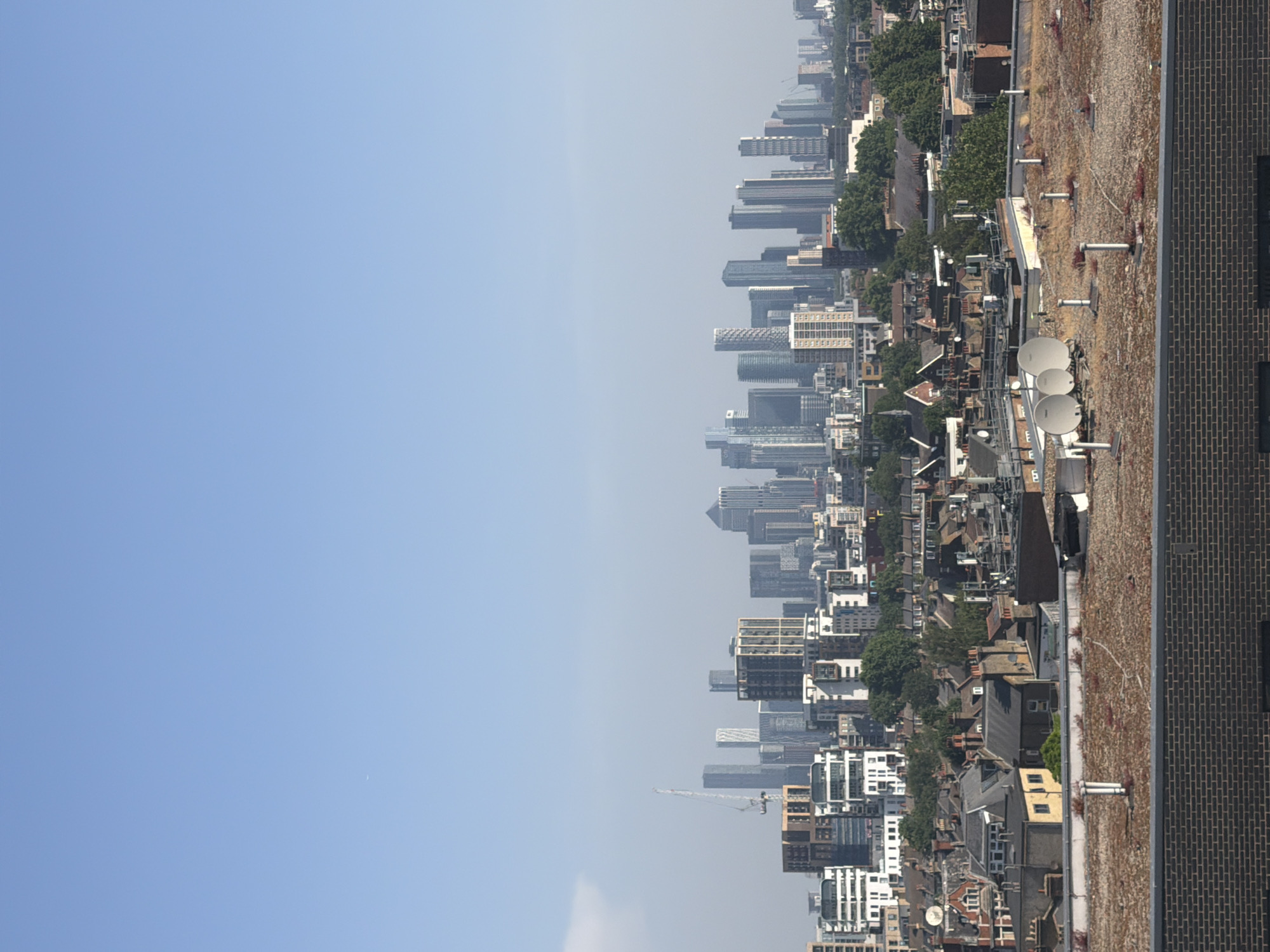 The image shows a view of London's skyline from a high vantage point, likely a rooftop. The sky is a clear, pale blue. Skyscrapers are visible in the distance, defining the city's modern skyline. A dense mix of residential buildings, interspersed with trees, fills the foreground, indicating a more urban area. Satellite dishes are mounted on several rooftops. A brick wall and a gravel-covered roof occupy the immediate foreground, suggesting the location is on a building's rooftop. Canary Wharf is a probable location given the distinctive skyscrapers. There are no people visible.