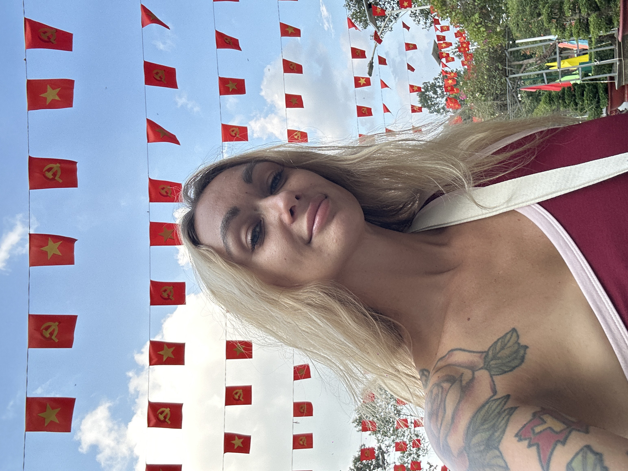 The image is a low-angle selfie of a fair-skinned woman with blonde hair. She is wearing a maroon top with white straps, and a floral tattoo is visible on her left arm. The background features strings of red flags with yellow stars and hammer-and-sickle symbols, suggesting a socialist or communist theme. The presence of these flags, commonly associated with Vietnam, indicates the photo was likely taken in the city of Ho Chi Minh City, Vietnam. A glimpse of trees and sky is visible between the flags.