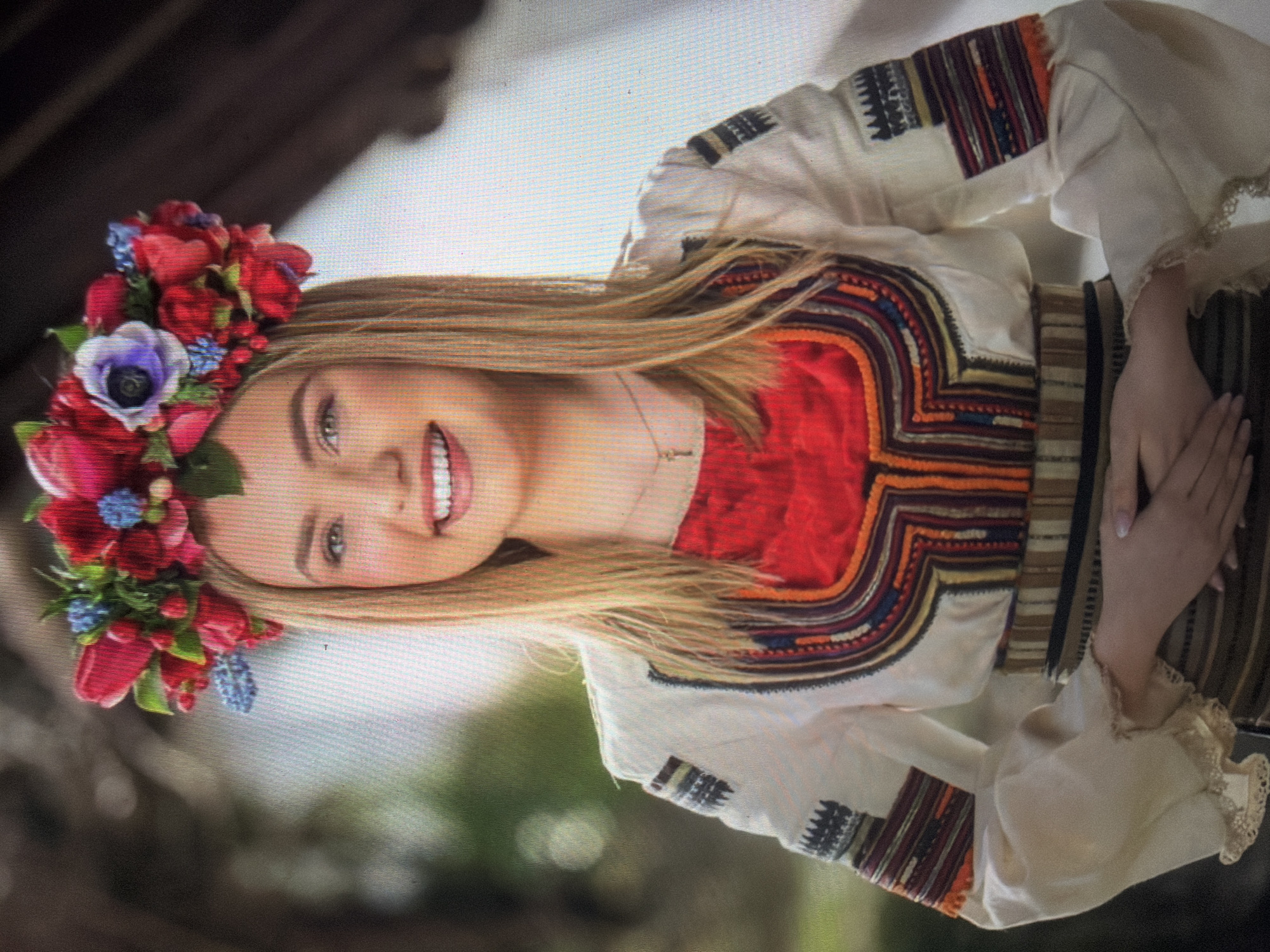 A young adult female is depicted in a half-body portrait, facing forward with a direct gaze and an open-mouth smile. She has long, straight, blonde hair parted in the center, and light-colored eyes. She wears a vibrant floral crown on her head, composed of numerous red tulip-like flowers, blue grape hyacinths, small red berries, green foliage, and a prominent purple and white anemone positioned above her forehead. Her attire consists of a traditional white long-sleeved blouse, featuring extensive multi-colored embroidery in geometric patterns across the chest, shoulders, and arm cuffs. The embroidery colors include red, orange, dark blue, black, and white. A red fabric panel is visible at the neckline of the blouse. The sleeves are voluminous and terminate in gathered, light-colored lace-trimmed cuffs. A horizontally striped woven sash in shades of brown and black is cinched at her waist. A delicate gold-toned chain necklace with a small, elongated pendant is visible around her neck. Her hands are gently clasped in front of her at waist level. The background is blurred, showing green foliage on the left side and darker, indistinct architectural elements, possibly wooden beams, on the right. This scene is located in the city of Shumen, Bulgaria.