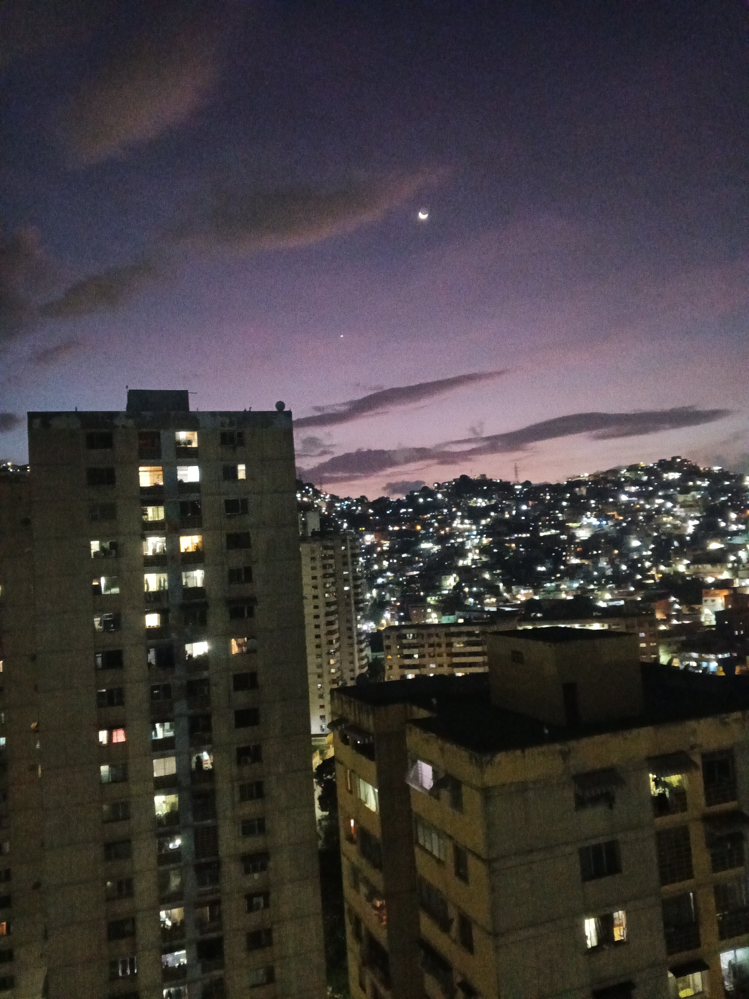 The image depicts a night cityscape under a twilight sky with a crescent moon. Tall buildings with lit windows are visible in the foreground, while a densely populated hillside with numerous lights stretches into the background. The overall impression is one of urban density and nocturnal activity.