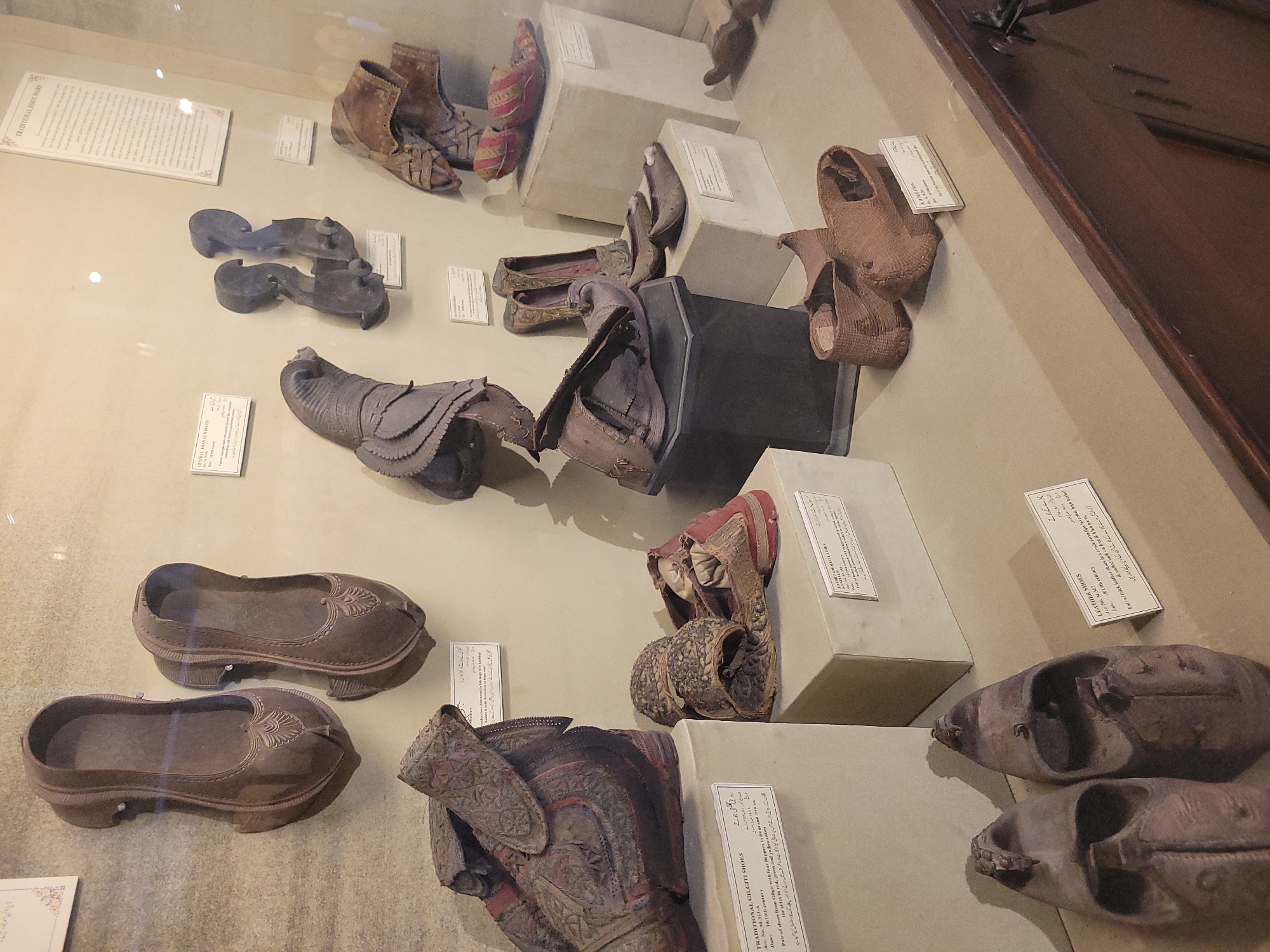 The image shows a museum display case filled with various traditional shoes and footwear. The display is lit from above, casting shadows on the objects. The background is a textured wall, possibly stone or plaster, in a neutral beige color.
The footwear items are presented on different pedestals and stands within the case. Some are arranged in pairs, while others are displayed individually. The types of footwear vary, including what appear to be leather shoes, embroidered slippers, and possibly clogs or sandals. Many of the items are ornate and appear to be made from leather, fabric, and possibly metal embellishments. Some pieces show signs of age and wear.
Labels are visible in front of some of the displayed items, providing descriptive text and possibly identification numbers or dates. These labels are white with black text.
In the foreground, the edge of a wooden display counter or cabinet is visible, with some metal hardware. The overall impression is that of a museum exhibit dedicated to historical or cultural footwear.