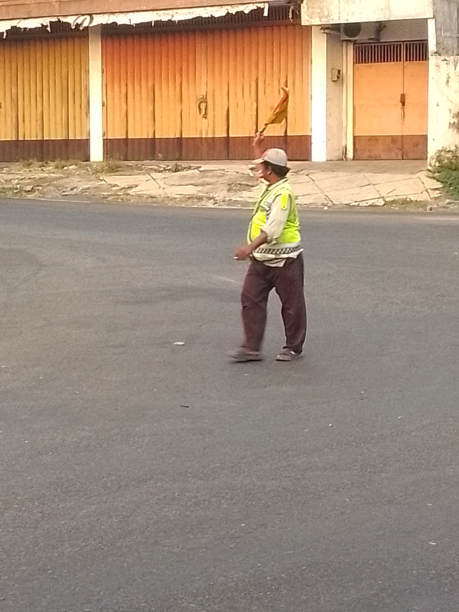 A full-body, slightly blurry, eye-level shot captures a middle-aged man wearing a light-colored long-sleeved shirt, a neon yellow safety vest, a baseball cap, and dark brown pants. He is walking from left to right across a paved road. He is holding a triangular, orange and yellow flag attached to a pole in his right hand, extending it behind him. The man has a light brown complexion and appears to be wearing sandals. Behind him, a row of closed, corrugated metal storefront gates with yellow and orange hues runs horizontally across the background. A white pillar separates two sections of the gates. To the right of the gates, a building facade is visible, featuring a cream-colored wall and a closed metal door. The scene is illuminated by daylight. This appears to be a street scene in Jember, Indonesia, possibly in a commercial area.