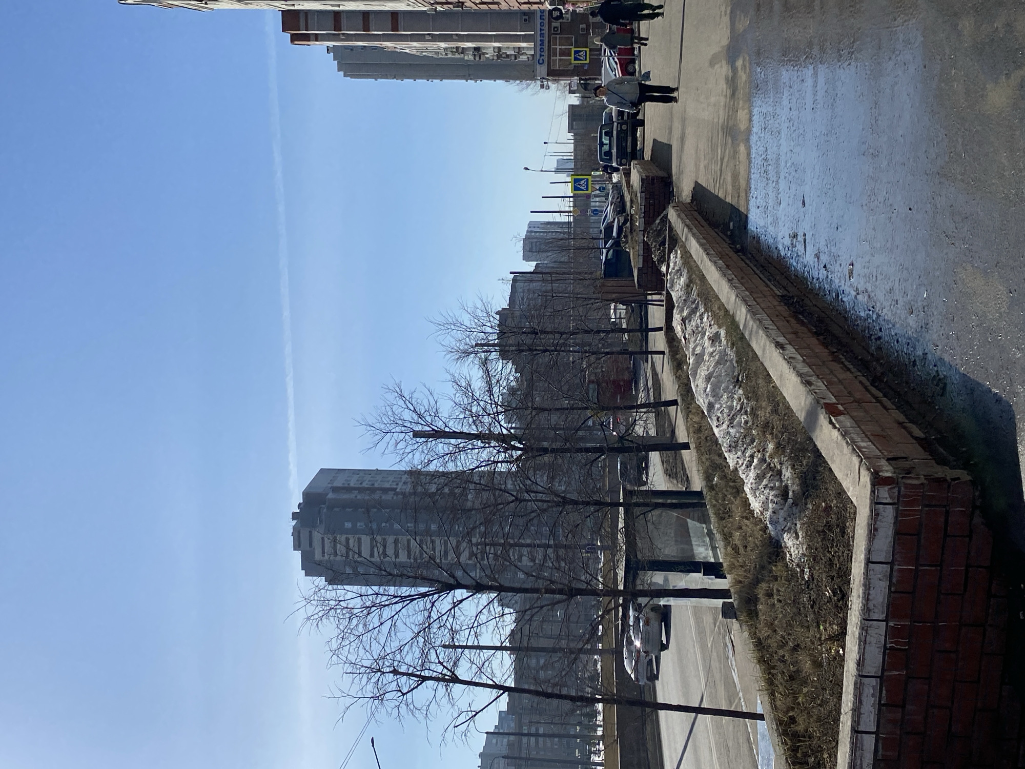 The image shows a street scene in Kazan, Russia. The sky is clear blue with a contrail. Tall apartment buildings line the street, bare trees in the foreground. A brick-edged flower bed runs alongside the wet pavement, with patches of snow. Cars are parked along the street. People are walking on the sidewalk. The weather appears to be cold. A sign with "СТОМАТОЛОГ" (Stomatolog, meaning Dentist) is visible on a building. A zebra crossing pedestrian sign is also present.