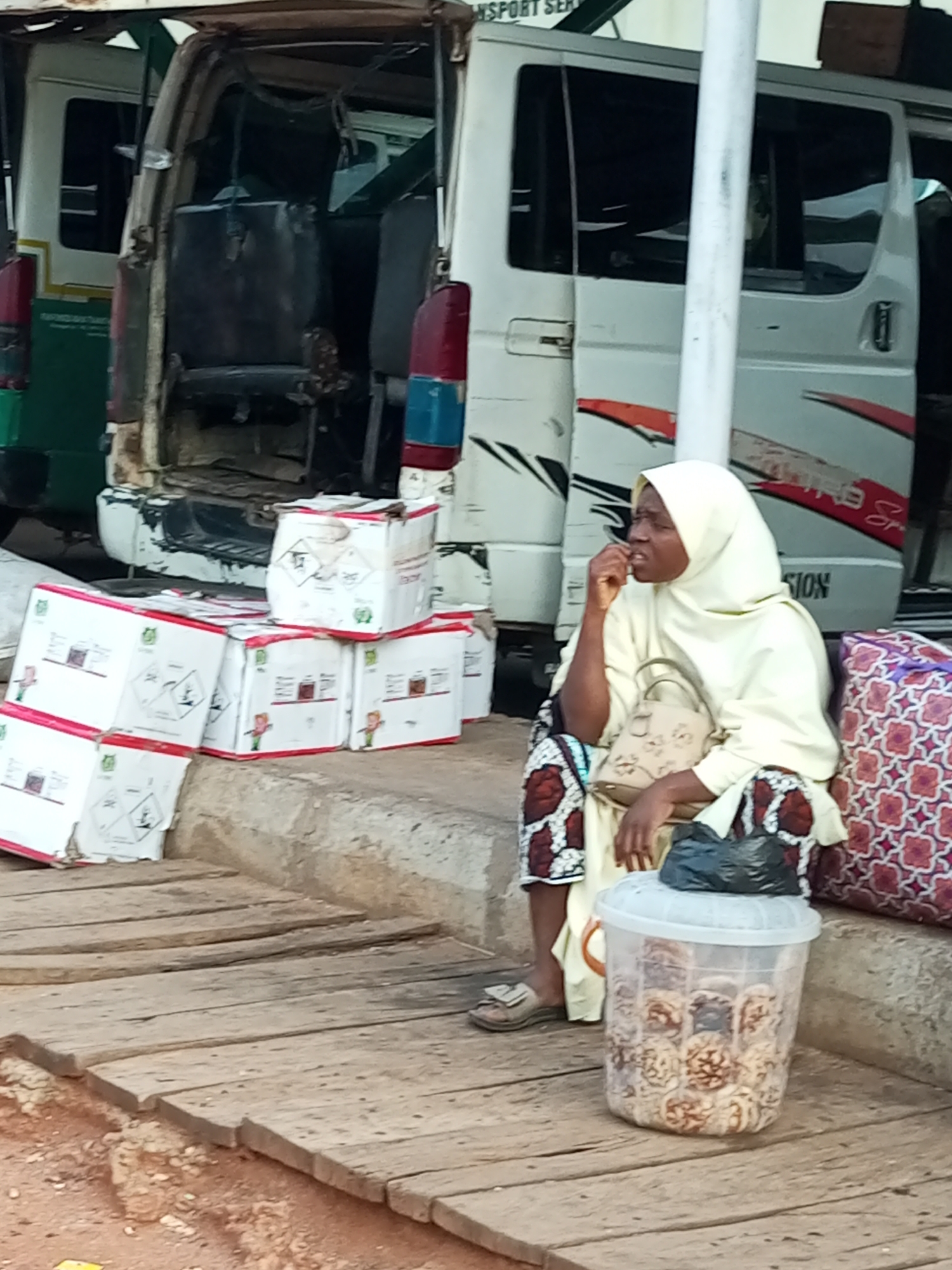 In Gombe, Nigeria, a woman wearing a yellow hijab and a patterned skirt sits on a wooden platform next to a white transport vehicle. She is positioned on the right side of the image, with her left hand near her mouth, appearing to be in thought or conversation. In front of her is a transparent plastic container filled with circular, light brown food items, possibly snacks or baked goods, topped with a black plastic bag. To her left, stacked white cardboard boxes with red trim are visible, some displaying hazard symbols and product images. The background shows the interior of the transport vehicle, with worn seats and debris, suggesting it is either out of service or being used for storage. The overall scene depicts a busy roadside or market area with elements of commerce and transit.