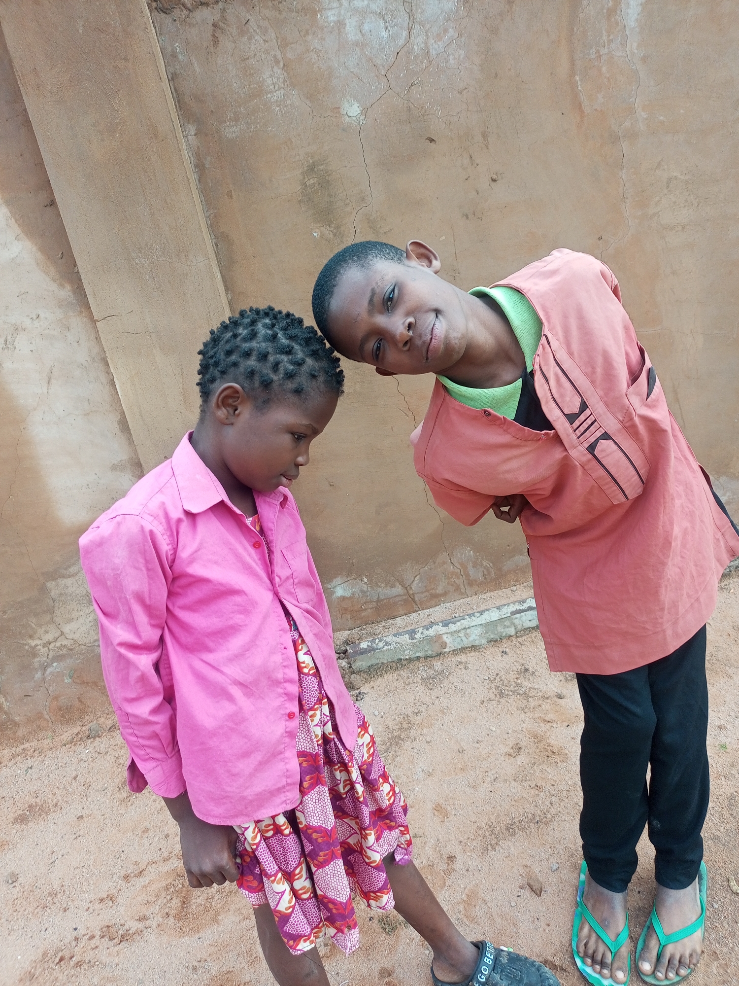 The image shows two children standing on dirt ground in front of a textured, light brown wall. The child on the left, appearing to be a girl, is wearing a bright pink collared shirt and a patterned skirt. Her hair is styled in short braids. She is looking down and slightly to her right. The child on the right, appearing to be a boy, is wearing a light green shirt underneath a coral-colored tunic with decorative black and red stitching. He has black pants and green flip-flops. He is leaning in towards the other child with his head tilted, smiling at the camera. The environment suggests an outdoor setting, possibly in a village or rural area within Tempure, Nigeria, given the simple construction of the wall and the unpaved ground.