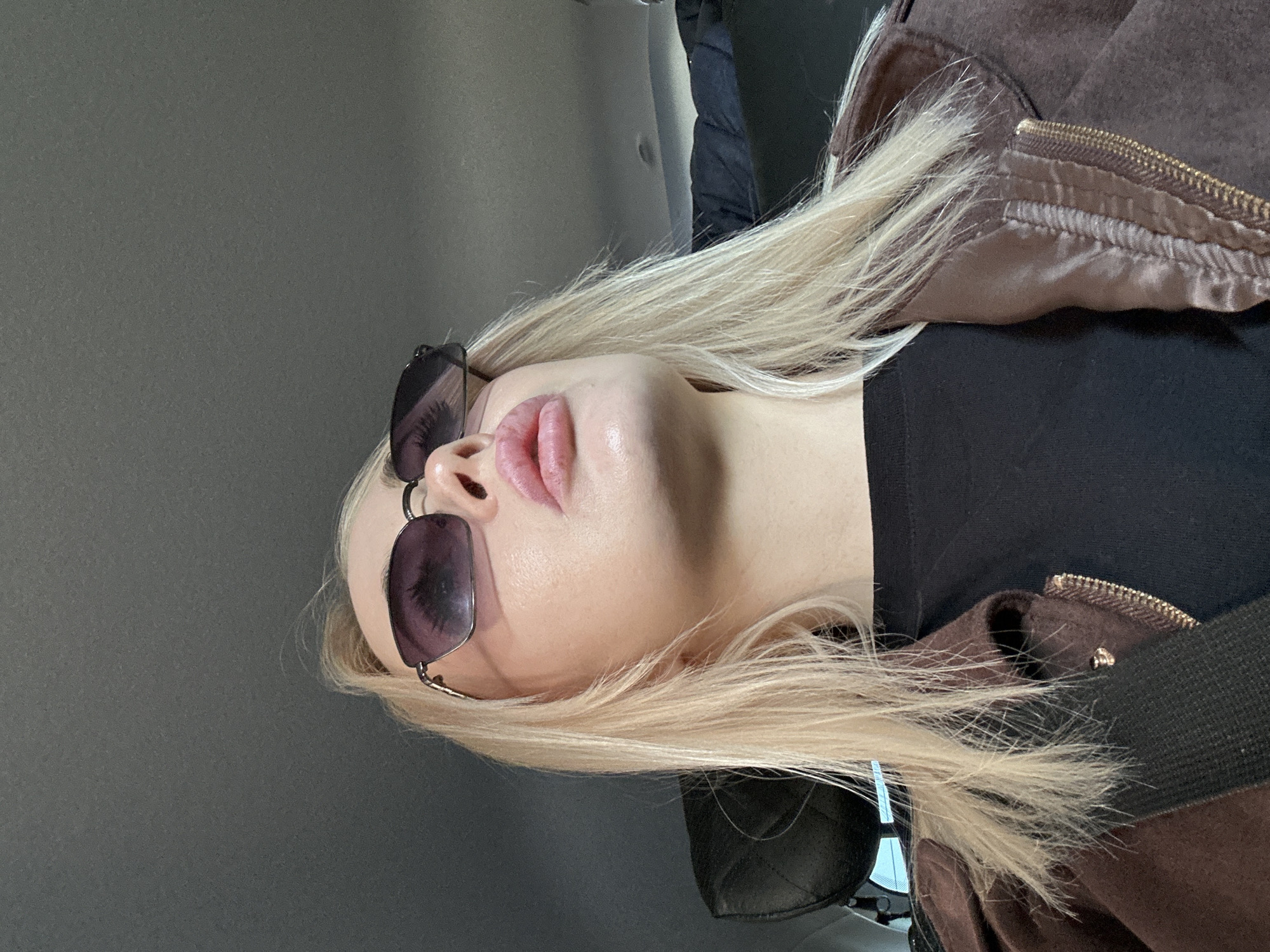The image shows a person with light blonde hair wearing sunglasses and a brown jacket. The background appears to be a grey wall. There is no indication that the photo was taken in Megion, Russia or any other specific location.