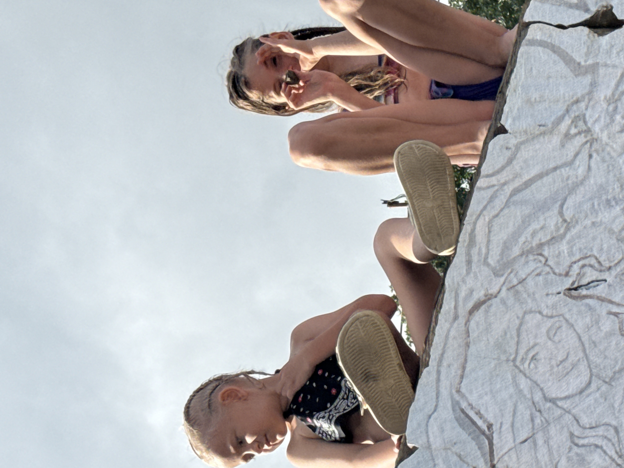 The image shows two young girls positioned on a large, textured surface that appears to be a rock or carved stone. The perspective is unusual, as if the viewer is looking up at them from below. Both girls are wearing casual clothing, with one in a swimsuit and the other in a bandana around her neck. One girl is holding something up to her eye, possibly a leaf or a stone, while the other girl is looking down. Their expressions are difficult to discern due to the angle and lighting, but they seem engaged in some playful activity. The background is a bright, overcast sky, suggesting it might be daytime, but the weather is unclear. The surface they are on has a carved relief of a face with flowing hair. The scene is outdoors, with hints of greenery in the background suggesting a natural or park-like setting. The overall impression is one of childhood play and curiosity.