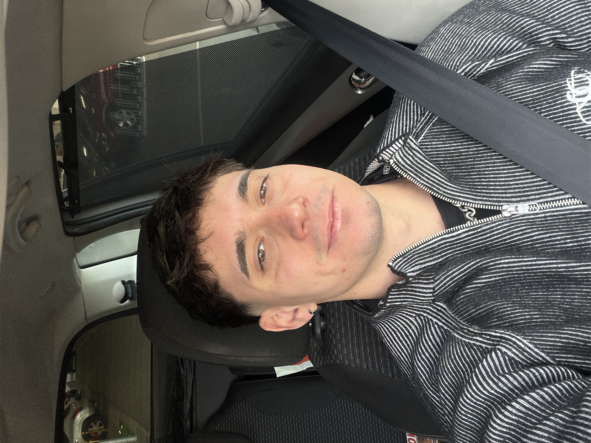 A young adult male, appearing in his 20s or early 30s, is centered in the frame, taking a selfie from inside a vehicle. He has short, dark brown hair with faded sides, brown eyes, a light complexion, and a faint smile. A small silver hoop earring is visible in his left ear. He is wearing a black and white vertically striped zip-up jacket over a dark undershirt, with a silver-toned chain visible. A black seatbelt diagonally crosses his chest, securely fastened. He is seated in the driver's seat, indicated by the seatbelt's position and the interior layout. The car's interior features dark grey/black textured fabric on the seat and light-colored ceiling and pillar trim. Through the vehicle's rear windows, the blurred outlines of other parked cars, including a silver SUV and a red vehicle, are discernible, suggesting a parking lot or garage environment. A mesh sunshade is partially visible on the right-side rear window. The scene is located within the city of Sofia, Bulgaria.