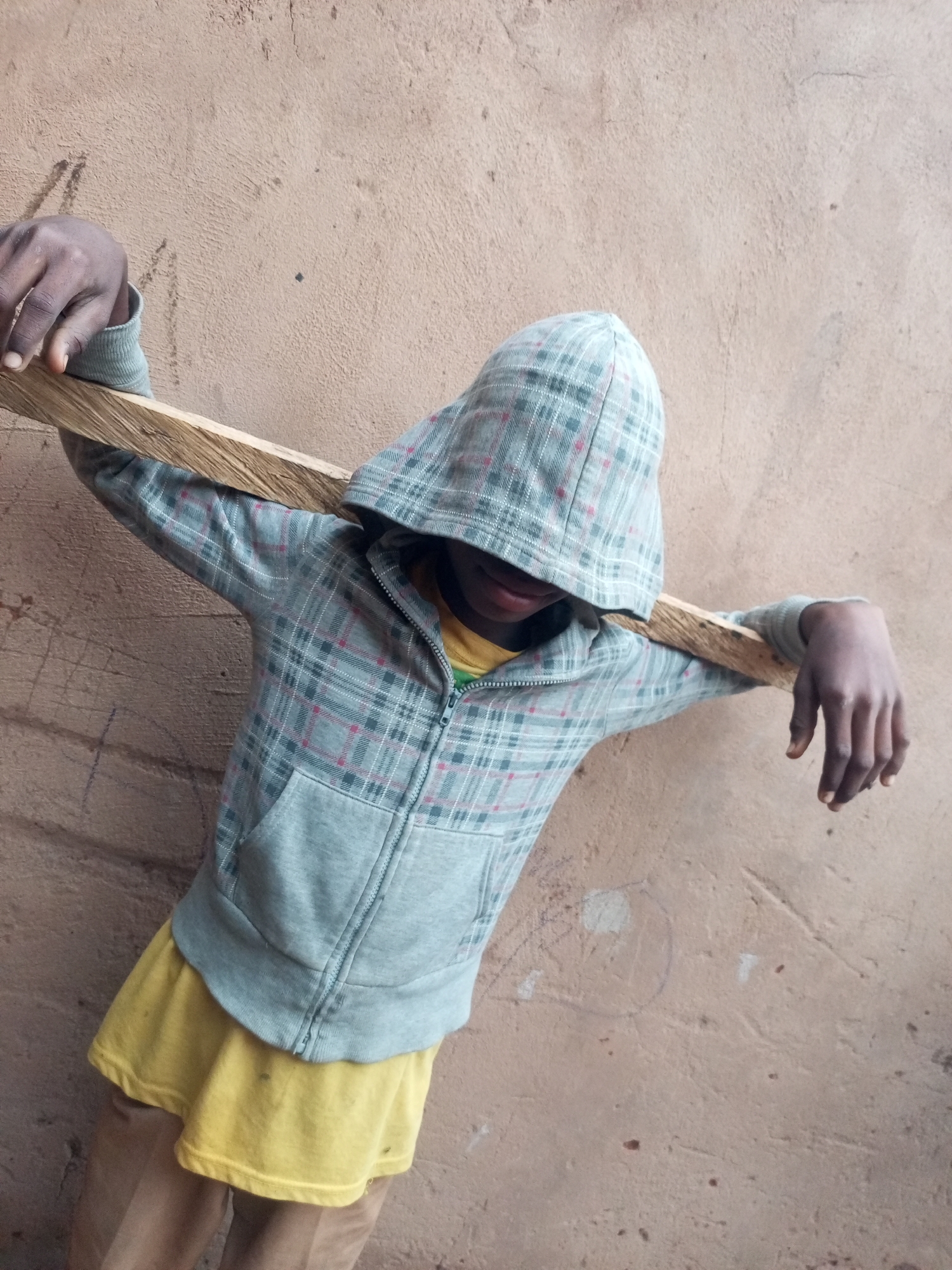A person wearing a grey plaid hooded jacket over a yellow t-shirt and light brown pants leans against a textured, reddish-brown wall. The person holds a light brown wooden plank horizontally across their chest, with their left arm resting on top of it and their right arm bent downwards, hand near their hip. The person's face is obscured by the hood. The background wall appears to be outdoors, possibly a street or exterior building wall, with some faint markings or scratches on its surface. The lighting suggests it is daytime. The image's overall tone is candid and unposed. The location is identified as Tempure, Nigeria.