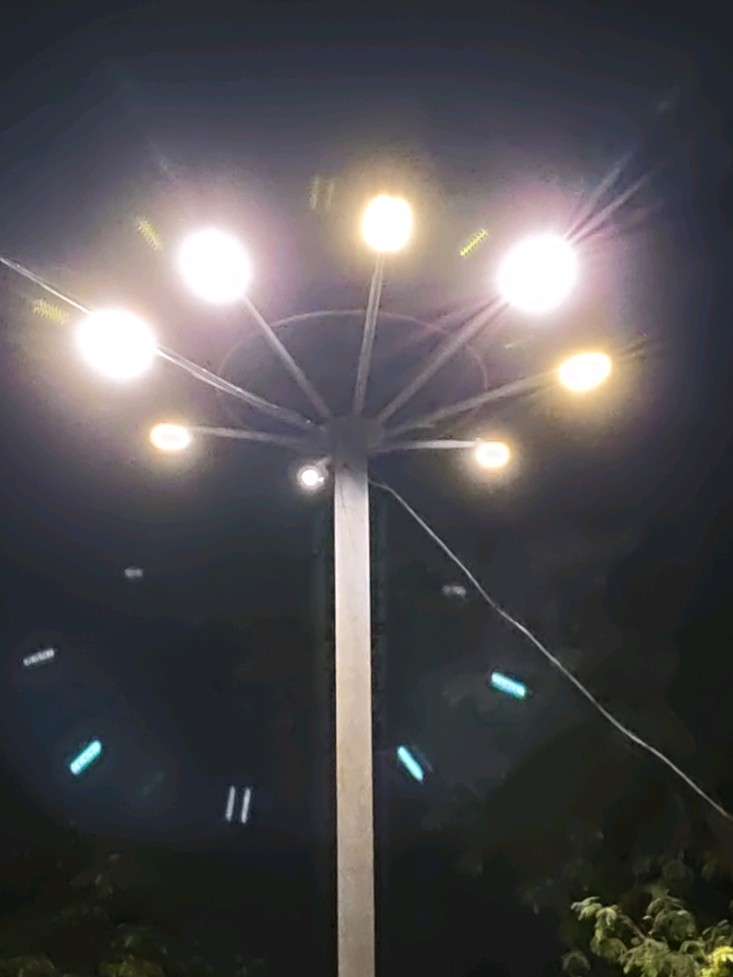 A tall lamppost is shown at night, with multiple lights emanating from the top. The lamppost is a thick, light-colored pole, with an umbrella-like structure at the top from which several lights extend. There are at least eight lights visible. Some of the lights are bright white, while others are a yellowish-white. There are some faint streaks of light and possibly some reflections on the lens. In the lower part of the image, some dark foliage is visible, suggesting the lamppost is outdoors, possibly in a park or along a street. There are also some small, glowing blue and white lights in the background, which could be other streetlights or decorative lights. The sky is dark, indicating nighttime.