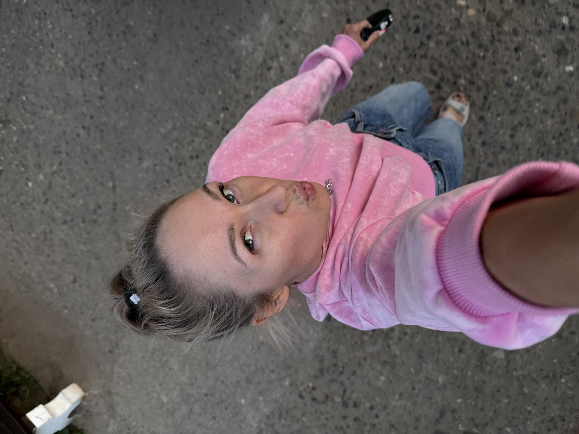 A woman stands on a gray paved surface, likely a sidewalk or street, and takes a selfie from a low angle, looking up into the camera. She wears a pink hooded sweatshirt and blue jeans and has blonde hair pulled back. She holds a small, dark-colored object in her right hand above her head, possibly a phone or remote control. She is wearing sandals. No specific location within Chaikovsky, Russia, can be determined from the image.