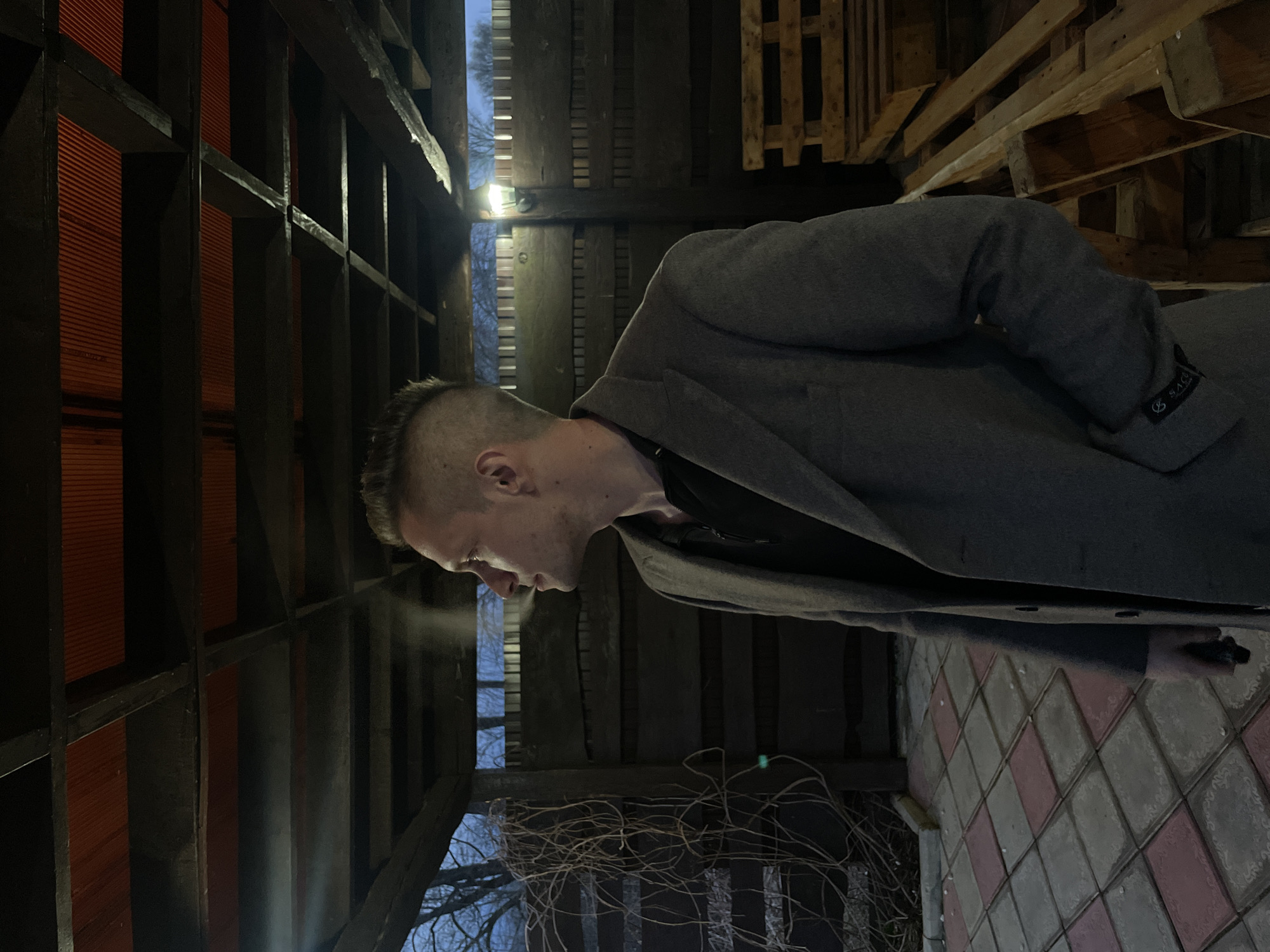 The media file displays a man in profile, standing outdoors in what appears to be a cold evening or night setting. He is positioned slightly right of center, looking downwards with his head tilted. The man has a short, faded haircut and is wearing a dark top under a long, grey coat. A small black tag with white text, which appears to say "SAGO," is visible on the cuff of his coat sleeve. His breath is visibly condensing in the air, indicating cold weather.
The setting appears to be an outdoor space within a wooden structure. Dark wooden beams frame the background, with reddish-orange corrugated panels visible through them on the left side of the frame. To the man's right, several wooden pallets are stacked. Bare tree branches are visible against a dark blue sky through gaps in the wooden structure. A light fixture is mounted on the wooden framework to the left of the man's head, casting some illumination. The ground is paved with diamond-patterned tiles in alternating shades of red/pink and grey/white. The overall scene suggests a quiet, possibly contemplative moment in a cool, semi-enclosed outdoor area.