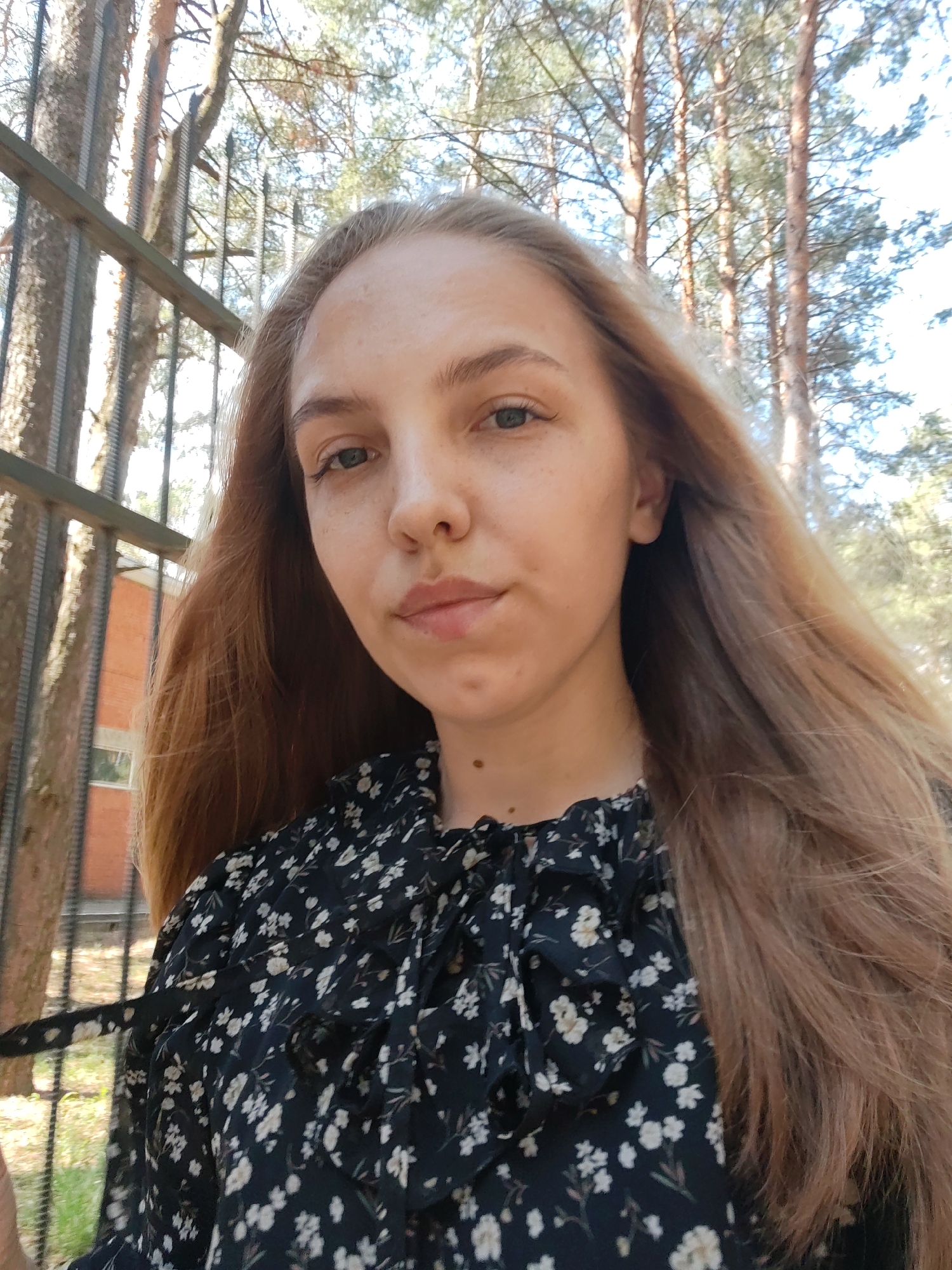 A young woman with long, light brown hair is pictured in a medium close-up shot outdoors, likely in Chaikovsky, Russia. She has fair skin, light eyes, and a slightly upturned nose. She wears a black floral dress with white flower patterns and a ruffled collar. Her hair is parted on the left side.
Behind her is a metal fence, a brick building with a visible window, and a wooded area with tall pine trees. The lighting indicates it is daytime, likely during the spring or summer.