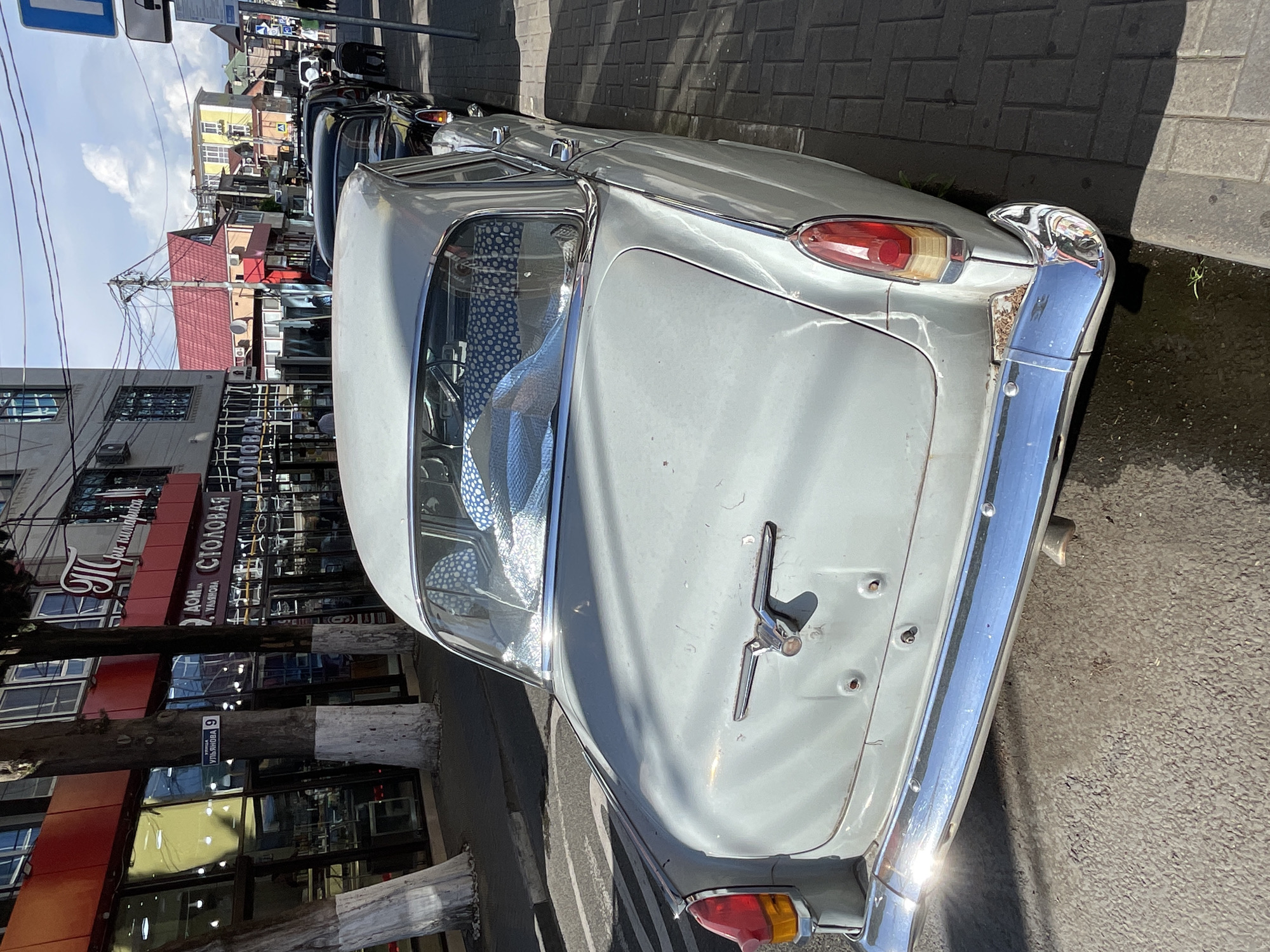 In Sochi, Russia, a light blue vintage sedan, likely a GAZ-21 "Volga", is parked on a street beside a brick sidewalk. The car is seen from a rear-quarter angle, highlighting its chrome bumper, taillights, and a distinctive eagle hood ornament on the trunk. The interior of the car, visible through the rear window, is covered with a blue and white polka-dot fabric. In the background, buildings line the street, with shops displaying signage in Russian. The sky is partly cloudy.