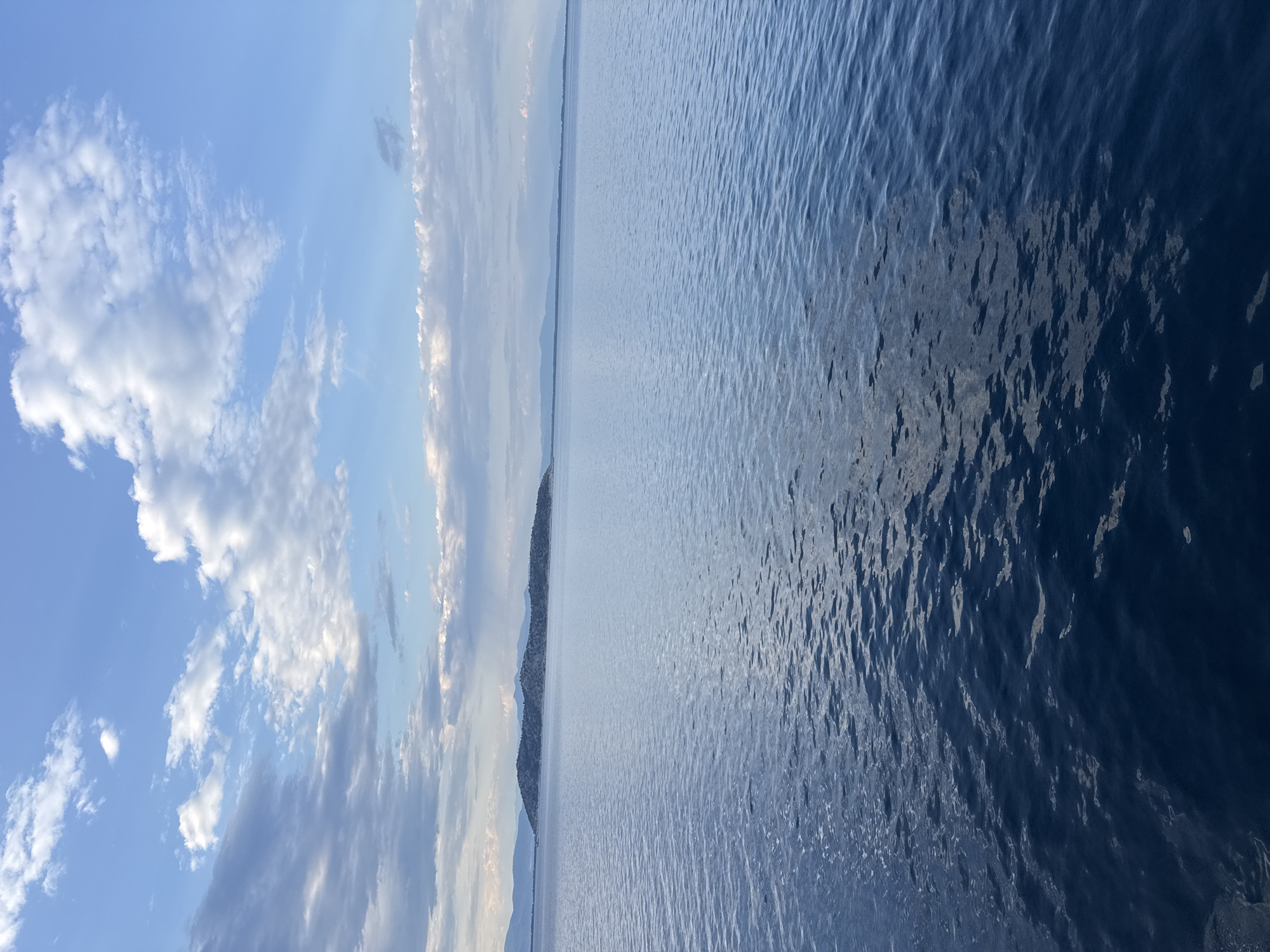 The image depicts a serene, expansive view of the Aegean Sea from an elevated vantage point, likely a boat or a high coastal area. The water is a deep, rich blue, with gentle ripples reflecting the sky and sunlight, creating a textured surface. In the middle distance, a long, low-lying island or peninsula is visible, characterized by a rugged, sparsely vegetated terrain with subtle mountainous features in the background. The coastline is indistinct and merges with the distant mountain ranges that form a hazy, muted horizon line. The sky above is a vibrant blue, adorned with scattered, fluffy white clouds that create dynamic patterns and add depth to the scene. The overall impression is one of tranquility and natural beauty, characteristic of the coastal landscapes of Thassos, Greece. No people, specific structures, or immediate actions are discernible.