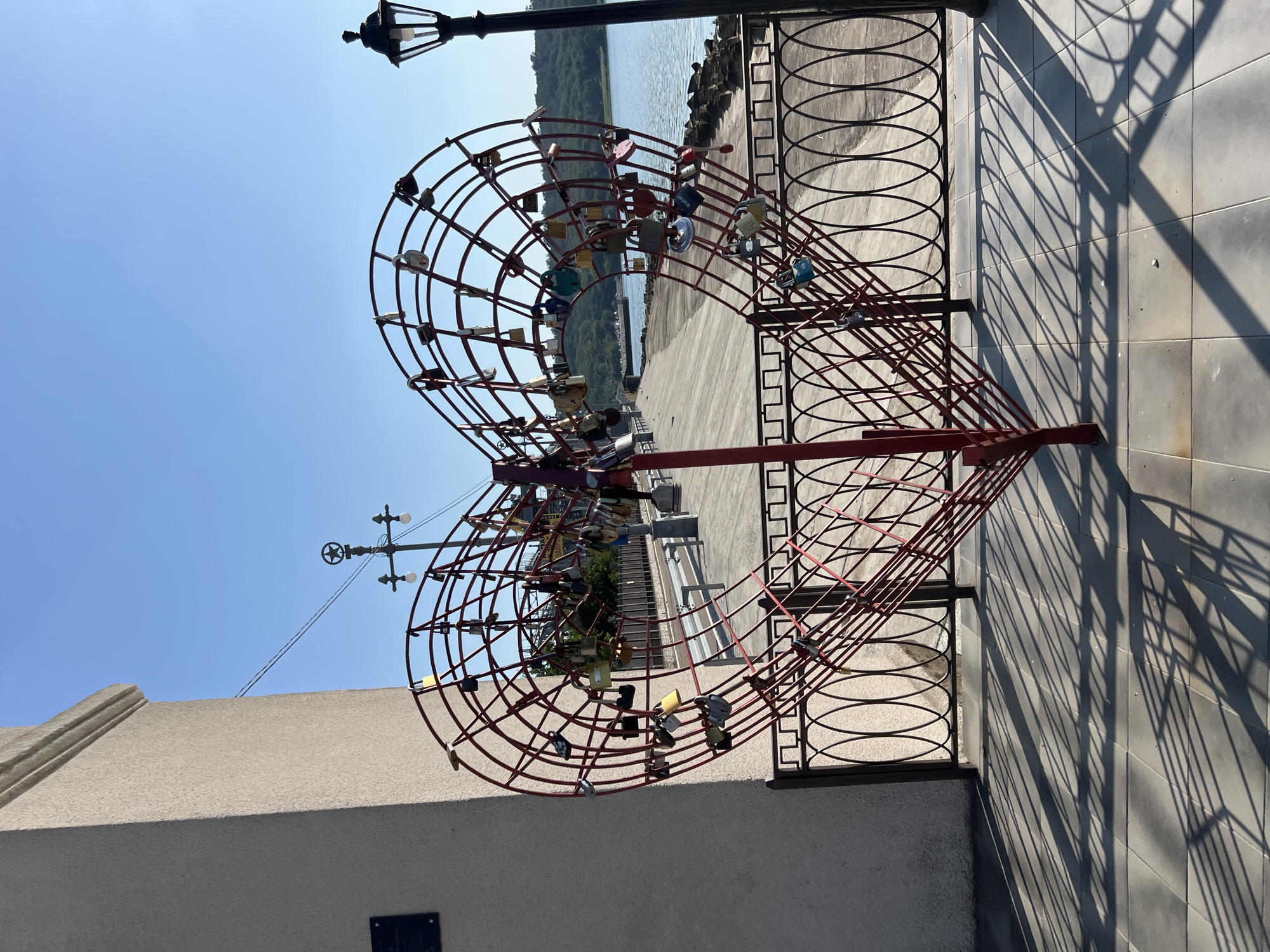 A large red metal heart sculpture, adorned with numerous padlocks, stands prominently in the foreground. The sculpture is located outdoors, adjacent to a promenade or walkway that runs alongside a body of water. To the left of the heart is a light-colored, textured wall of a building. A black metal fence runs in front of the sculpture and separates it from the promenade. A lamppost with a star on top is visible behind the sculpture, and another, more traditional lamppost is to the right. The background features a body of water, hills with trees, and a clear blue sky. The scene is illuminated by sunlight, casting long shadows from the sculpture and fence onto the tiled ground. The city of Korzhevo, Moldova is mentioned in the prompt.