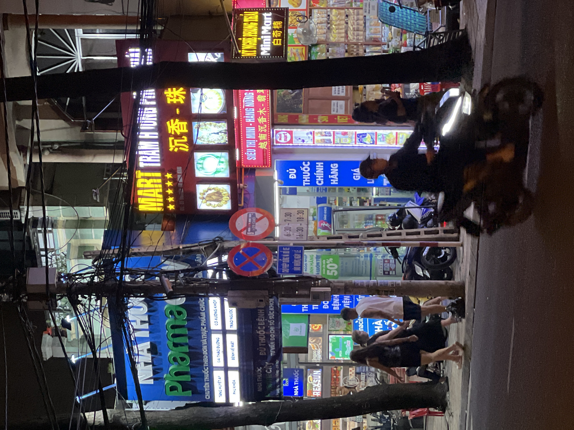 This is a nighttime street-level photograph taken in Nha Trang, Vietnam. The scene is dominated by a busy street with several illuminated signs for businesses. A large blue sign with green lettering for "Nhà Thuốc Pharmacy" is prominently displayed. Adjacent to it, a red sign with yellow lettering reads "MART TRẦM HƯƠNG". Further to the right, another red sign announces "SIÊU THỊ MINI - HÀNG NÔNG" in white and red lettering, with a "Mini Mart" sign below it. There are also multiple smaller signs in Vietnamese, including one for "REX SUN".
People are visible in the image: a group of three individuals are walking on the sidewalk on the left, and a person is riding a motorcycle in the foreground, moving from right to left across the frame. The environment includes trees, street poles with numerous wires, and parked motorcycles. The overall impression is a vibrant commercial street at night.