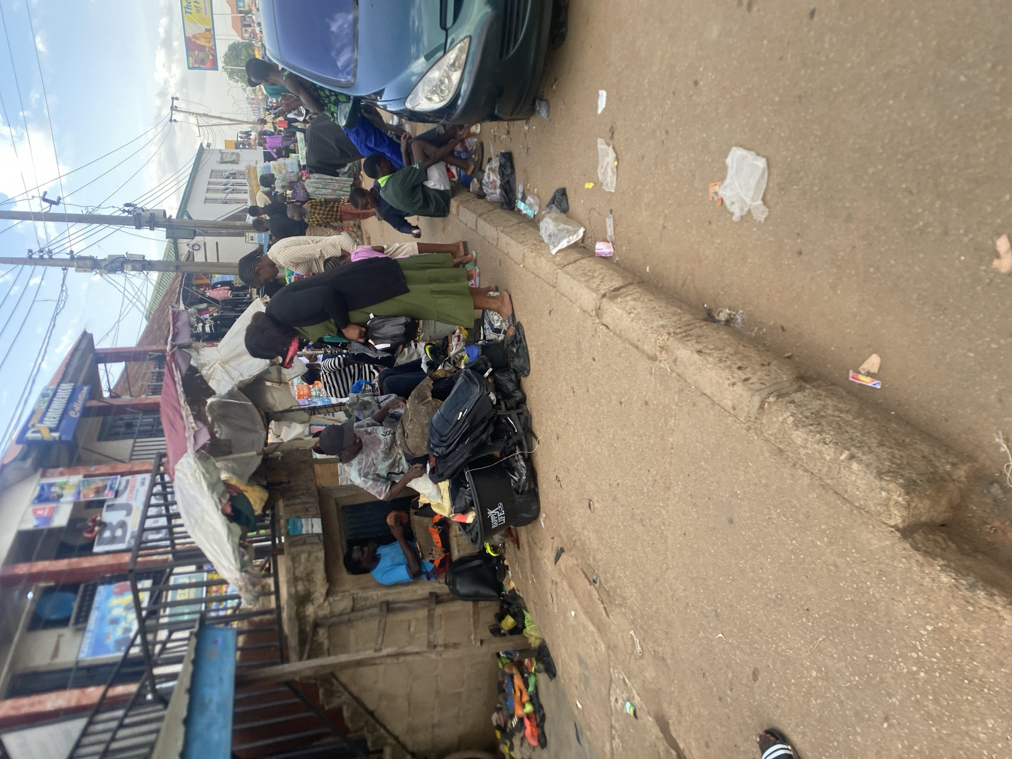 The image depicts a bustling street scene in Bukuru, Nigeria, showcasing a vibrant roadside market or commercial area during what appears to be daytime, under a partly cloudy sky.
Numerous individuals are visible, including vendors and customers, engaged in various activities. Many people are seen bending over or sitting among goods displayed directly on the ground along a concrete sidewalk. In the foreground, a woman wearing a green dress and dark top is focused on items laid out. Nearby, a man in a blue shirt is seated among merchandise. The goods include a variety of bags, shoes, and clothing. One black bag is clearly marked with the text "HAPPY LIFE."
A dark green car is partially visible on the paved road to the right, adjacent to the sidewalk. The ground both on the road and along the curb shows scattered litter, such as plastic bags and wrappers. In the background, several multi-story buildings line the street. One building features a blue sign that reads "PREMIUMCO Collection," with another sign partially visible below it displaying "BJ." Overhead electrical poles and wires crisscross against the sky, and a metal staircase is attached to one of the buildings, indicating a mixed-use commercial and residential environment. The scene portrays a lively everyday atmosphere of street commerce and pedestrian movement.