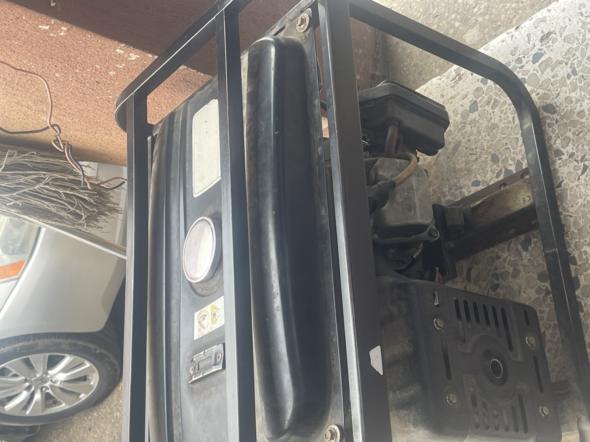 The image displays a black portable generator, appearing used and somewhat dusty, situated on a light-colored, patterned terrazzo-style floor. The generator is encased in a sturdy black metal frame. Its main body includes a prominent metallic fuel cap and a control panel area on the left side, while engine components like the air filter housing and muffler are visible on the right.
To the immediate left of the generator, the front fender and a multi-spoke wheel of a silver car are partially visible. Below the car, a pile of dry, fibrous material, resembling straw or broom bristles, is present. Above the generator, a light brown, textured surface, possibly a ceiling or an overhang, is shown, with loose orange/brown and black electrical wires hanging from it.
The scene suggests an outdoor or semi-outdoor utility or storage area, likely under bright daylight conditions, indicating a sunny day. No human activity or individuals are present.
Visible details on the generator include a circular warning symbol depicting a crossed-out lit cigarette, signifying "No Smoking" or "Flammable material." Below it, another warning symbol is partially visible, likely related to electrical safety. The letters "CE" are also faintly discernible on a sticker or plate on the lower right side of the generator, indicating European conformity. The overall impression is that of functional equipment in a practical setting, consistent with a utility space in Abuja, Nigeria.