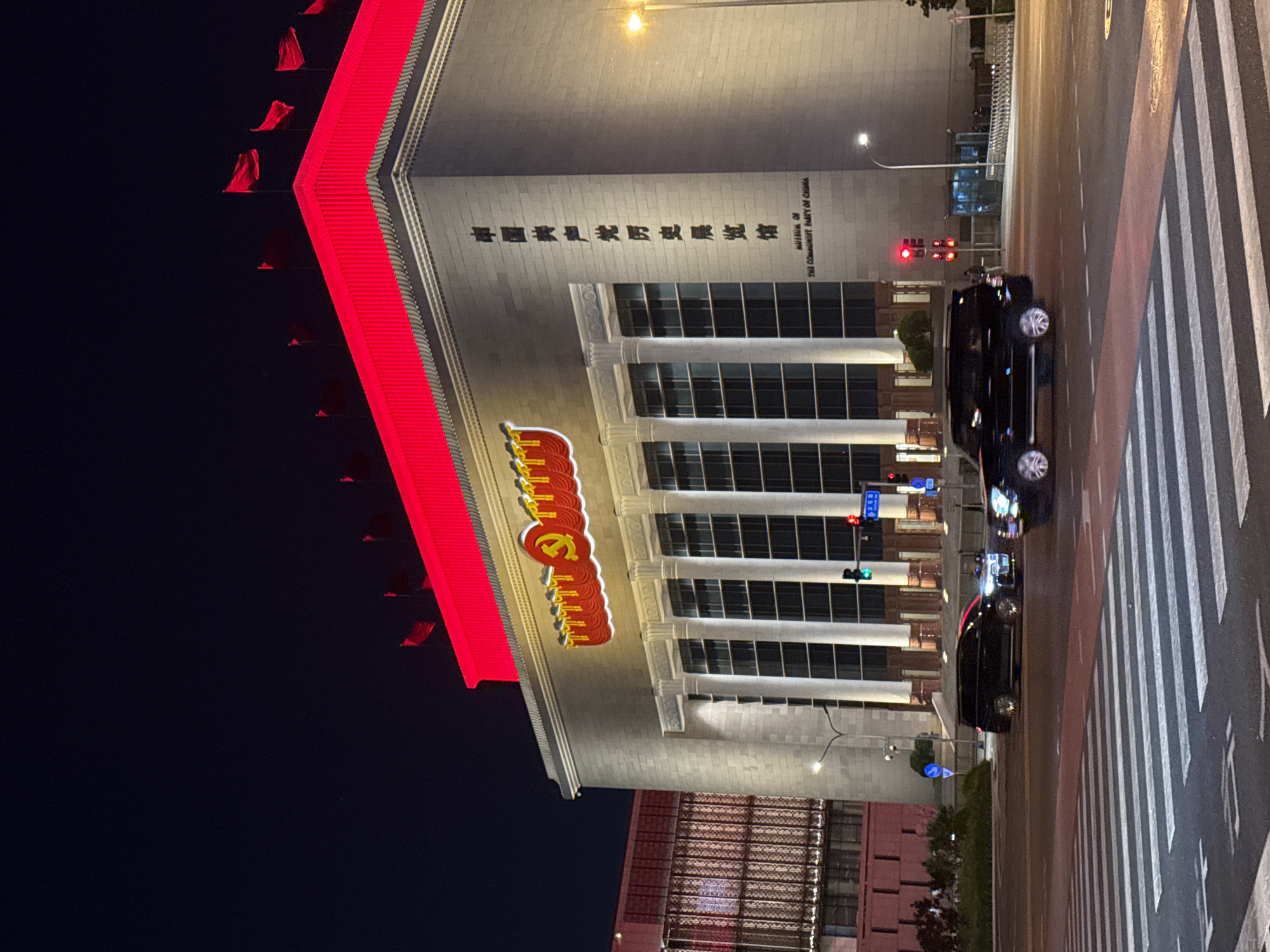 The image captures the Museum of the Communist Party of China at night in Chaoyang District, China. The building is illuminated, with its facade featuring the communist hammer and sickle emblem. A bright red banner adorned with multiple flags runs along the side of the building. In front, there is a crosswalk on a road with vehicles present. The sky is dark, contrasting with the brightly lit building and street.