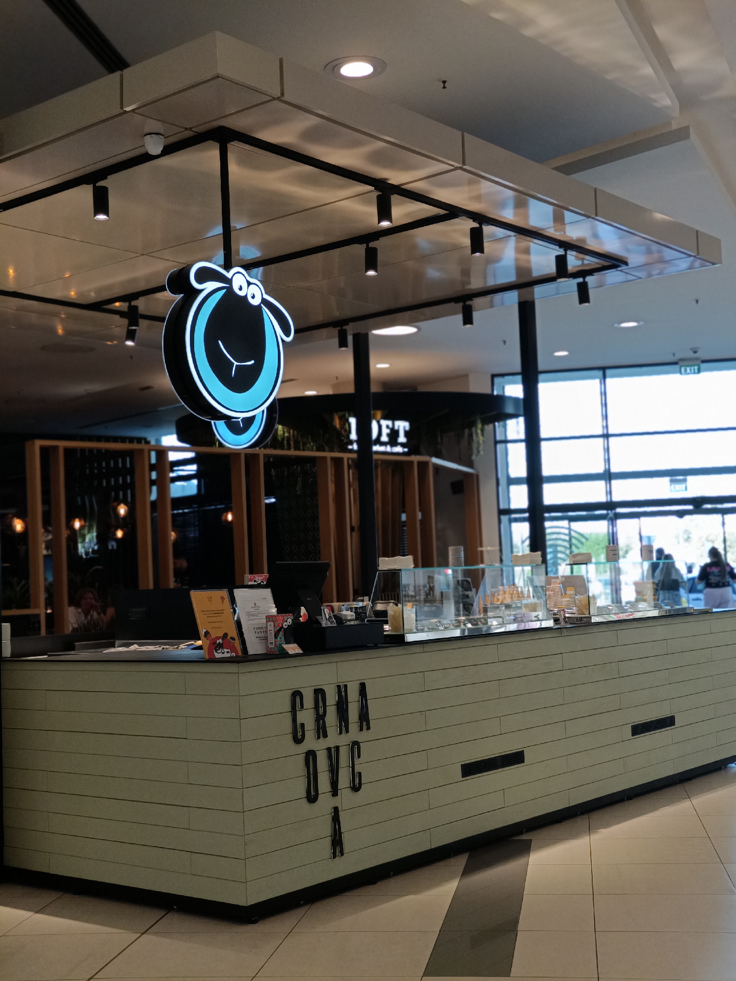 The image shows the interior of a commercial establishment, likely a cafe or dessert shop, in a modern shopping mall. The main subject is a counter area with a display case filled with food items, possibly ice cream or pastries. Above the counter, a distinctive logo featuring a stylized black sheep with bright blue accents is prominently displayed, illuminated by neon lights.
The counter itself is made of light-colored, horizontally striped material, with black lettering on the front that reads "CRNA OVC A," which translates to "Black Sheep" in Croatian or Serbian. There are also ventilation grates visible on the side of the counter. Behind the counter, a black cash register and various promotional materials are set up.
In the background, there's a glimpse of other parts of the establishment, including wooden paneling and lighting. Through large glass windows, the exterior of the building or another section of the mall is visible, with people walking by and an "EXIT" sign. The lighting suggests it is daytime, with artificial lights from the ceiling and the shop's own lighting contributing to the overall brightness. The scene appears clean and well-maintained, indicating a typical operational environment for a retail business. The overall impression is one of a welcoming and modern food service venue.