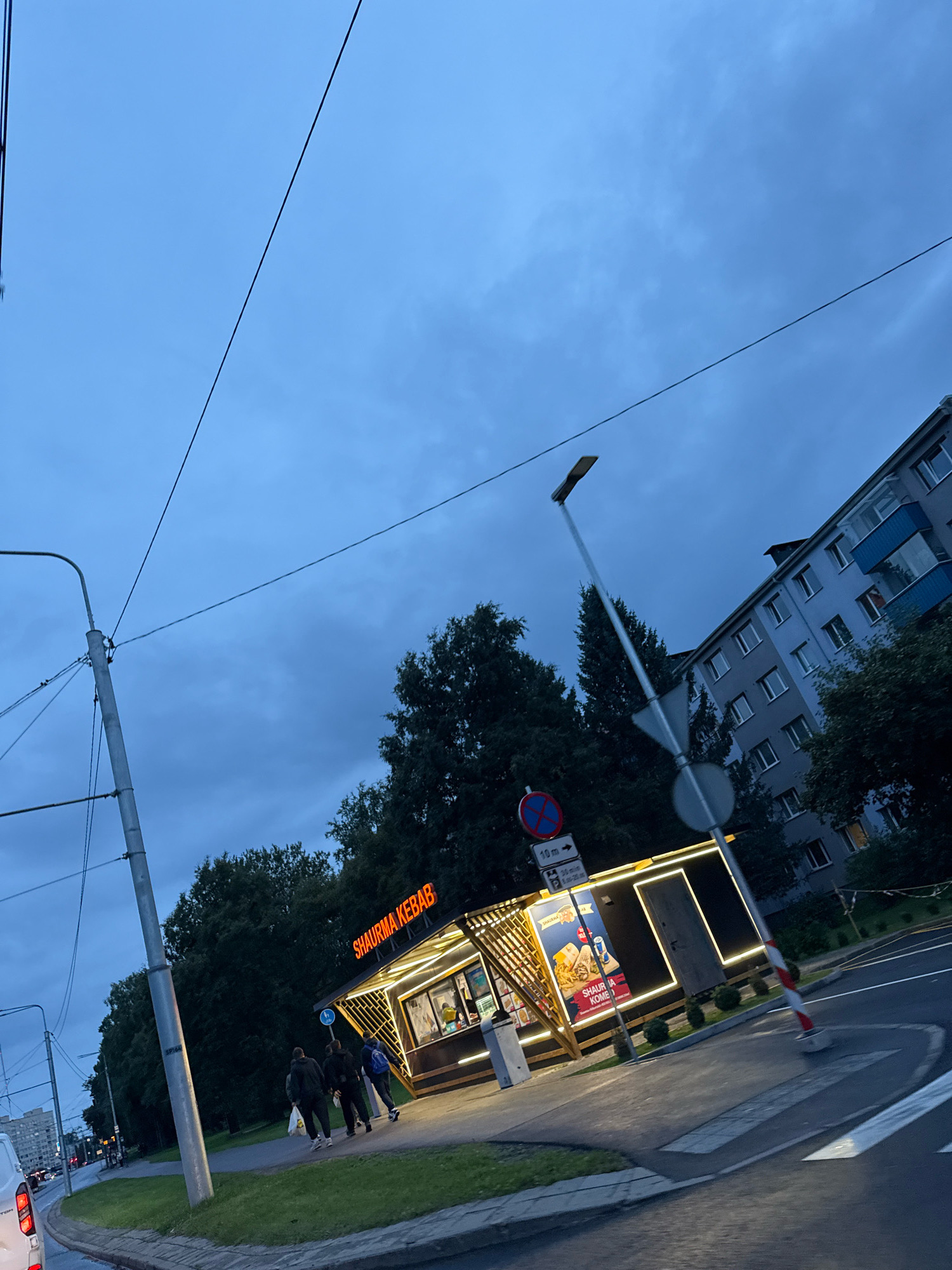 The image depicts a street scene at dusk in Tallinn, Estonia, likely in a residential or mixed-use area. The focal point is a brightly lit, modern shawarma stand with the sign "SHAURMA KEBAB" prominently displayed in orange lettering. The stand has a wooden exterior and is outlined with glowing white lights. It appears to be open for business, with a menu board visible on the side featuring pictures of food, including fries and what looks like a shawarma wrap. A few customers, dressed in casual attire, are walking away from the stand, one carrying a plastic bag.
In the background, a multi-story apartment building with light-colored facades and blue balconies is visible. Trees line the street, and overhead power lines crisscross the sky. A street lamp with a modern design is also present, along with various traffic signs, including a red and blue circular sign and a rectangular sign with parking information and a directional arrow. A pedestrian crossing is marked on the asphalt road in the foreground. The overall atmosphere suggests a typical urban evening in Tallinn, with the contrast between the illuminated food stand and the darkening sky.