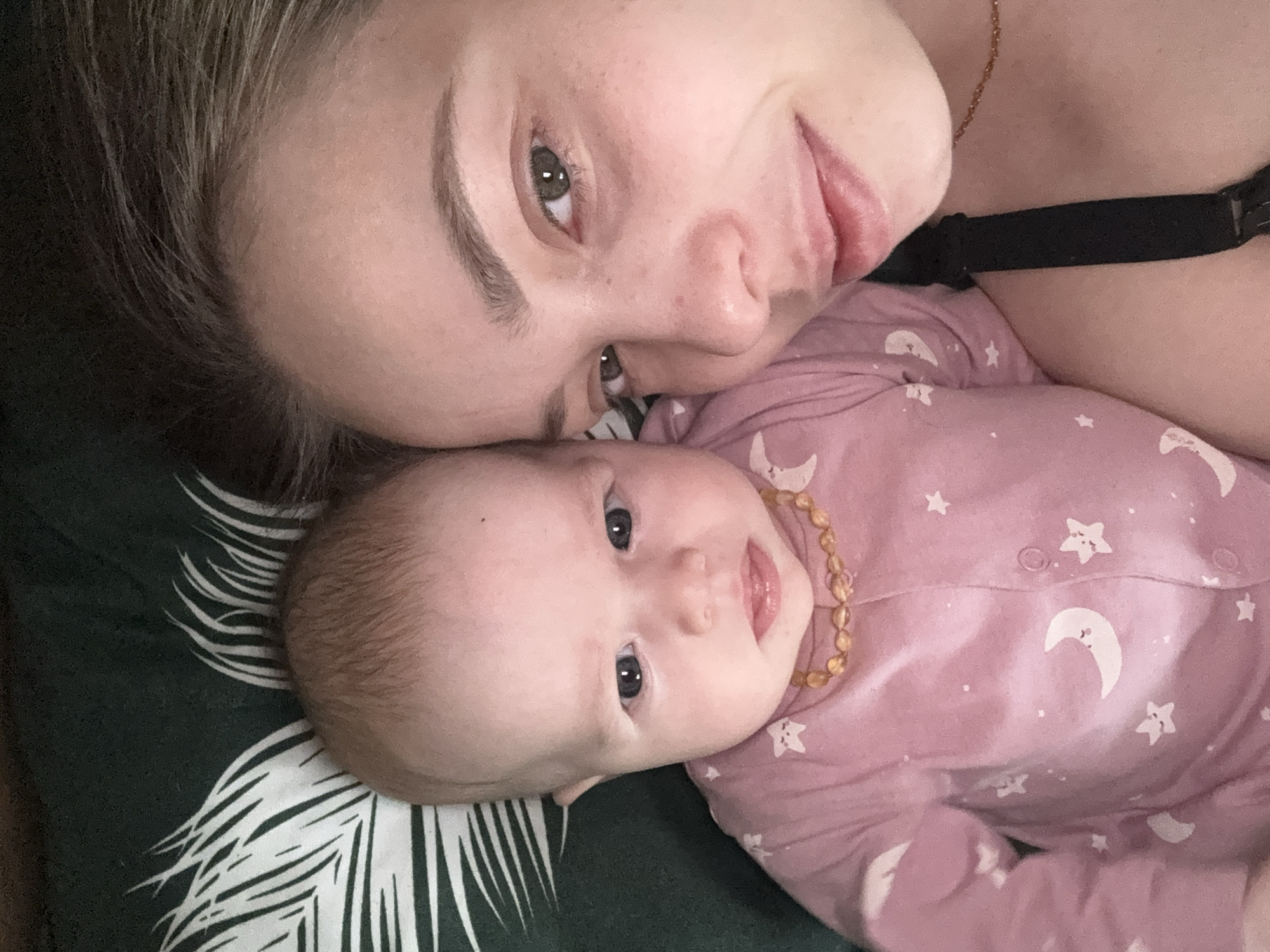 A close-up selfie shows a woman lying next to a baby on a patterned surface. The woman is light-skinned with green eyes and light brown hair. She is partially facing the camera, with her head resting on the surface. She is wearing a black bra strap. The baby, also light-skinned, is wearing a pink onesie with white star and moon patterns and a beaded necklace. The baby is looking towards the camera. The background is a green surface with a white palm leaf pattern. There is no indication of the location being Chaikovsky, Russia, or any city for that matter.