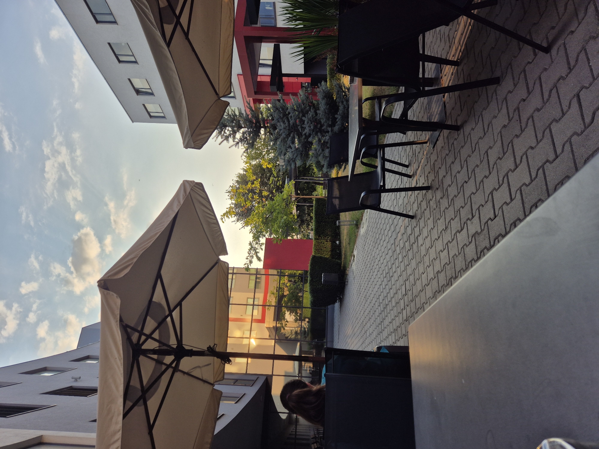 The image shows an outdoor seating area with tables and chairs under large umbrellas. In the foreground, a dark-colored table is partially visible. A paved walkway with interlocking gray bricks leads to a group of black metal chairs and tables, some of which are positioned near a small evergreen tree. Behind the seating area, there is a modern building with a glass facade reflecting the setting sun. The building features a red accent color on some of its architectural elements. Lush green bushes and trees are scattered around the area. A person with long brown hair is seated at a table on the left, facing away from the camera. The sky above is a mix of blue with scattered clouds, indicating it is late afternoon or early evening.