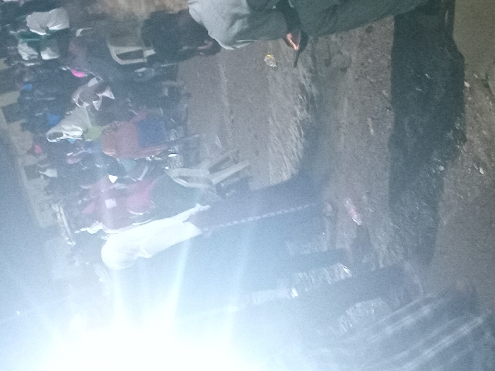 A dimly lit, blurry image shows a crowd of people congregated outdoors, potentially at night. Individuals are densely packed together, wearing various clothing. Some appear to be seated in chairs. A light source causes significant glare at the bottom of the image, obscuring details. The background appears to be a concrete or asphalt surface, with a wall running along one side. The overall impression is one of a gathering or event taking place in poor lighting conditions.