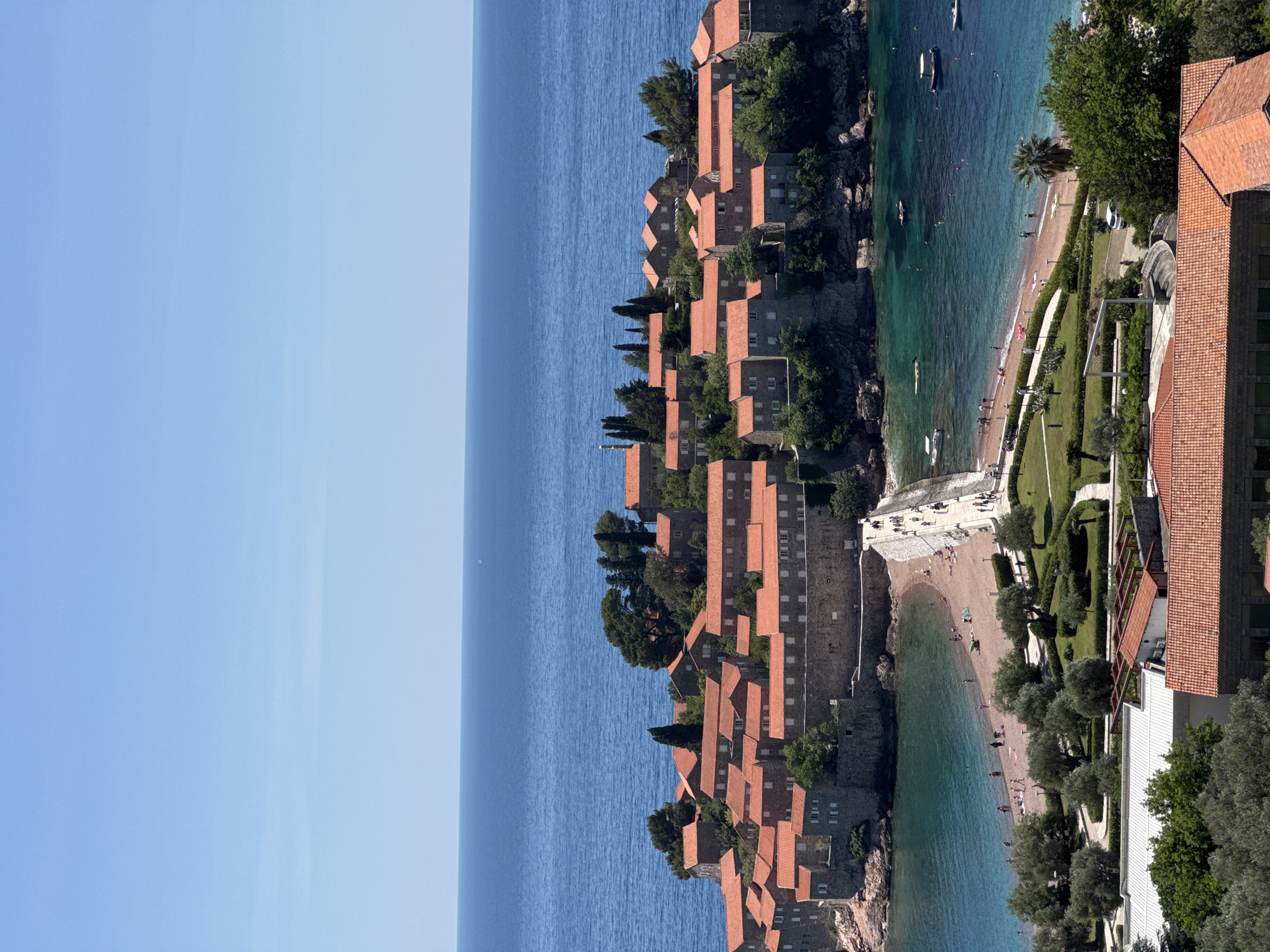 This aerial view depicts Sveti Stefan, Montenegro, a small islet connected to the mainland by a narrow isthmus. The islet is densely packed with closely arranged buildings, predominantly featuring red-tiled roofs. The buildings, likely residential or tourist accommodations, are positioned atop the rocky islet.
The islet is surrounded by turquoise water, with visible boats and people swimming. Two beaches flank the isthmus, one to the left and one to the right.
The mainland adjacent to the isthmus features manicured lawns, trees, and buildings. People are visible on the beaches, pathways, and grassy areas, some swimming, others walking.