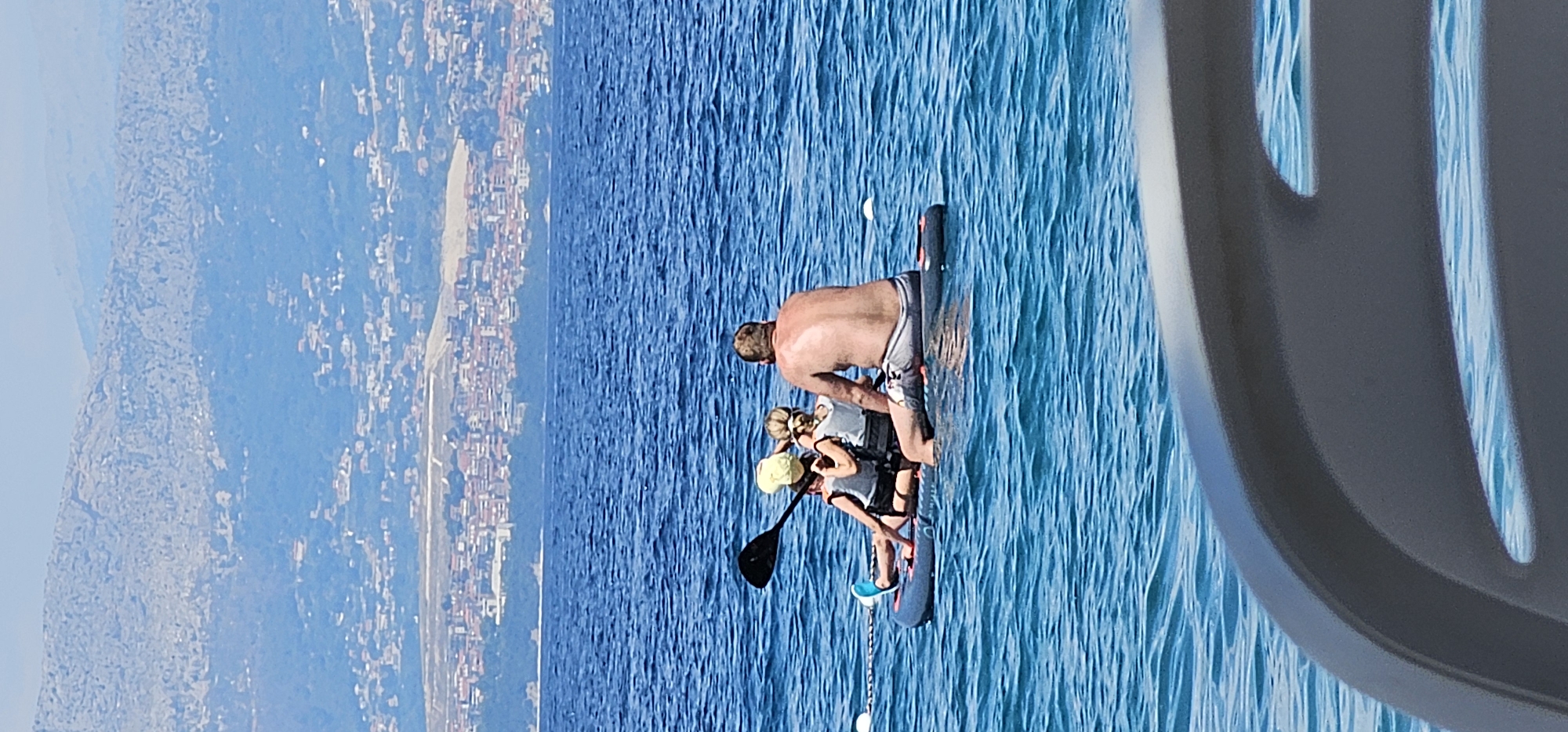 The image depicts a sunny day at sea, with clear blue water and a distant coastline. In the foreground, a paddleboard carries three individuals: a man with his back to the camera, his skin tanned and showing signs of sun exposure, wearing grey shorts. Two children, facing away from the viewer and towards the land, are seated on the paddleboard. One child is wearing a yellow hat and a life vest, while the other, slightly behind, also wears a life vest. The man is holding a paddle, suggesting they are engaged in paddleboarding. The paddleboard is a dark color with red accents. The sea is calm, with gentle ripples. In the background, the city of Supetar, Croatia, is visible, characterized by a densely packed collection of buildings with red-tiled roofs, nestled against rolling, sparsely vegetated hills that rise into mountainous terrain. The overall scene conveys a sense of leisure and outdoor activity on a warm day.