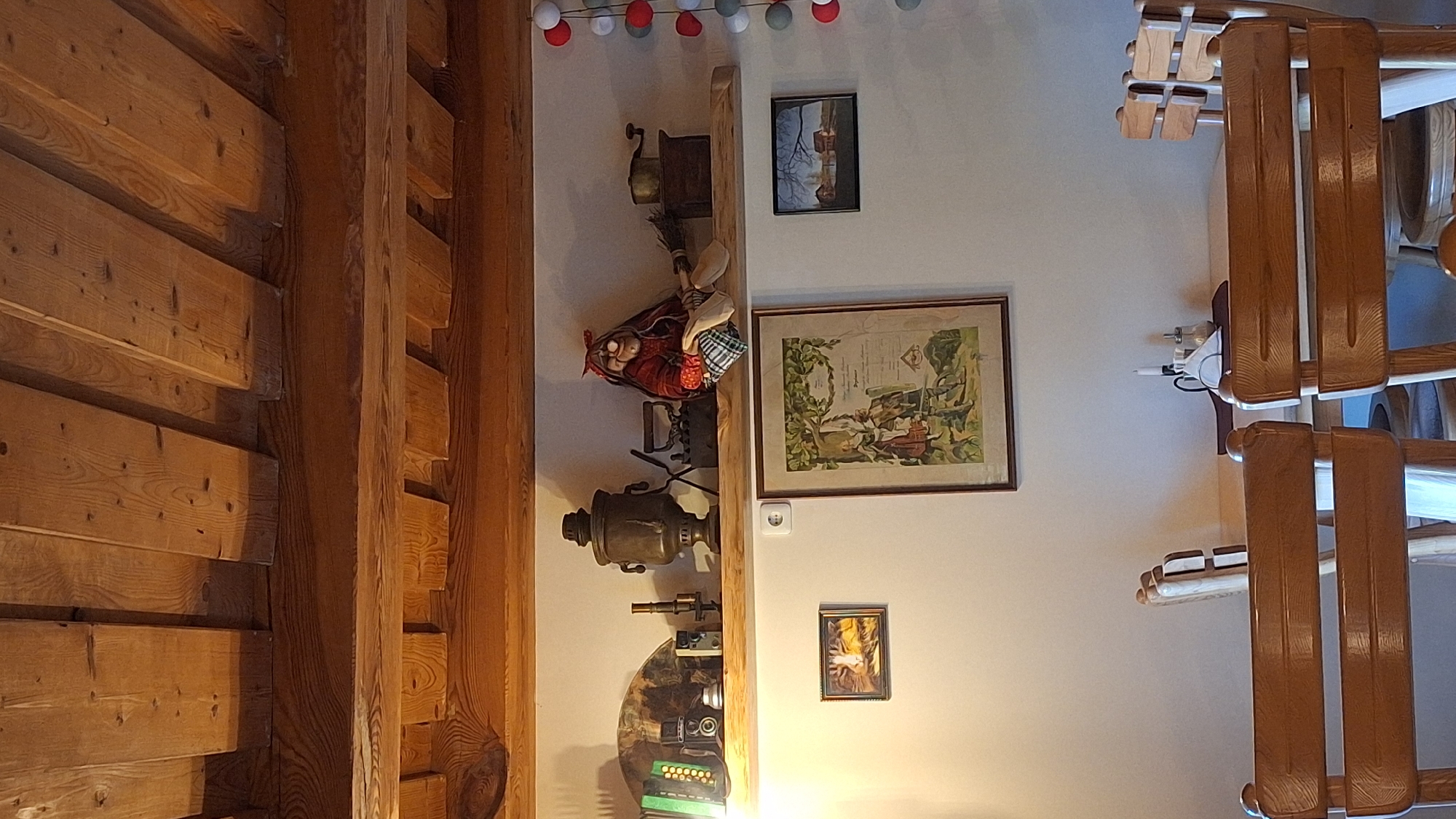 The image depicts a rustic interior scene, possibly a restaurant or dining area, featuring wooden elements throughout. A prominent wooden shelf is mounted on a light-colored wall, displaying various decorative items. These include what appears to be an accordion, cameras, a vintage samovar, two old-fashioned irons, a coffee grinder, and a doll dressed in traditional clothing. Below the shelf, two framed pictures are hung on the wall. The left picture is a close-up of a cat's face, and the larger central picture contains a colorful illustration with text. To the right of the central picture, a smaller landscape painting of a house by a body of water is visible. In the foreground, two wooden chairs with horizontal slats are positioned at a wooden table. On the table, a white napkin is neatly folded, accompanied by a salt and pepper shaker set and a candle in a holder. The ceiling is made of exposed wooden beams and planks, contributing to the rustic ambiance. The overall impression is one of a cozy and traditionally decorated space. Based on available information, this scene is located in the city of Lēdmane, Latvia.