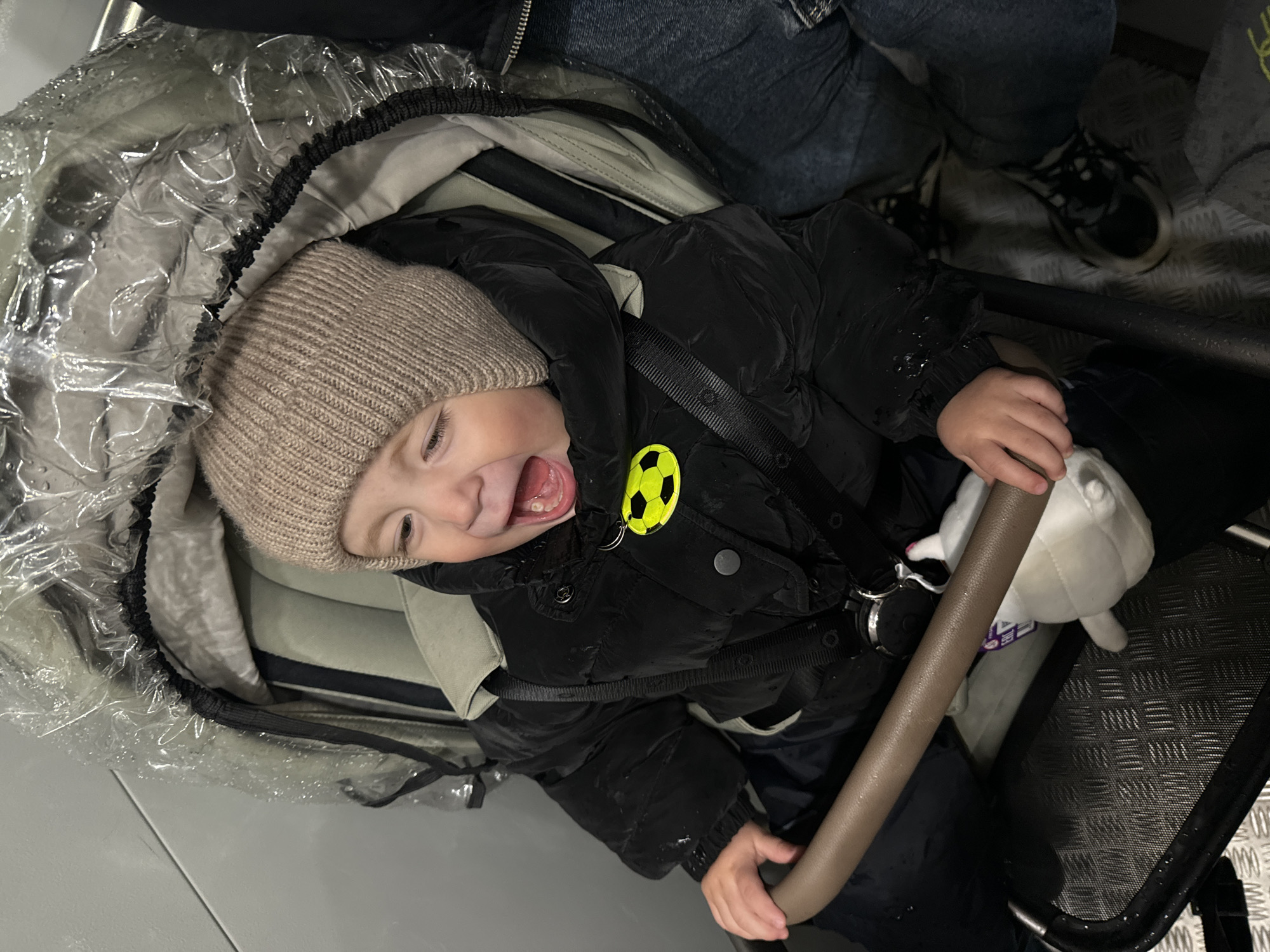A child wearing a tan knit cap and a black puffer jacket sits in a stroller with a clear plastic rain cover. The child is smiling with their mouth open. A stuffed animal is held in the child's hands. A person wearing jeans and sneakers is seen partially in the background. The scene appears to be indoors, possibly on public transportation. There is no information in the image to suggest this is in Odintsovo, Russia.