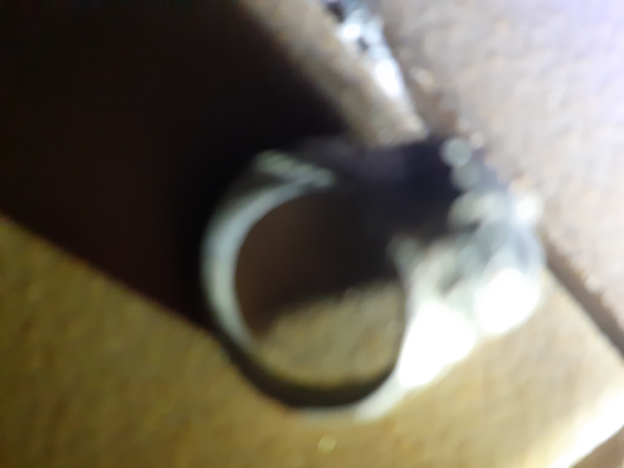 A close-up, out-of-focus image depicts a ring resting on a textured, wooden surface. The ring is silver-toned and appears to have some intricate detailing or etchings, though the blurriness obscures precise features. The background is dark and indistinct, with some lighter areas suggesting ambient lighting. The overall impression is of a personal item placed within a domestic or workshop setting. The location in Jalingo, Nigeria, is not discernible from the visual information provided in the image.