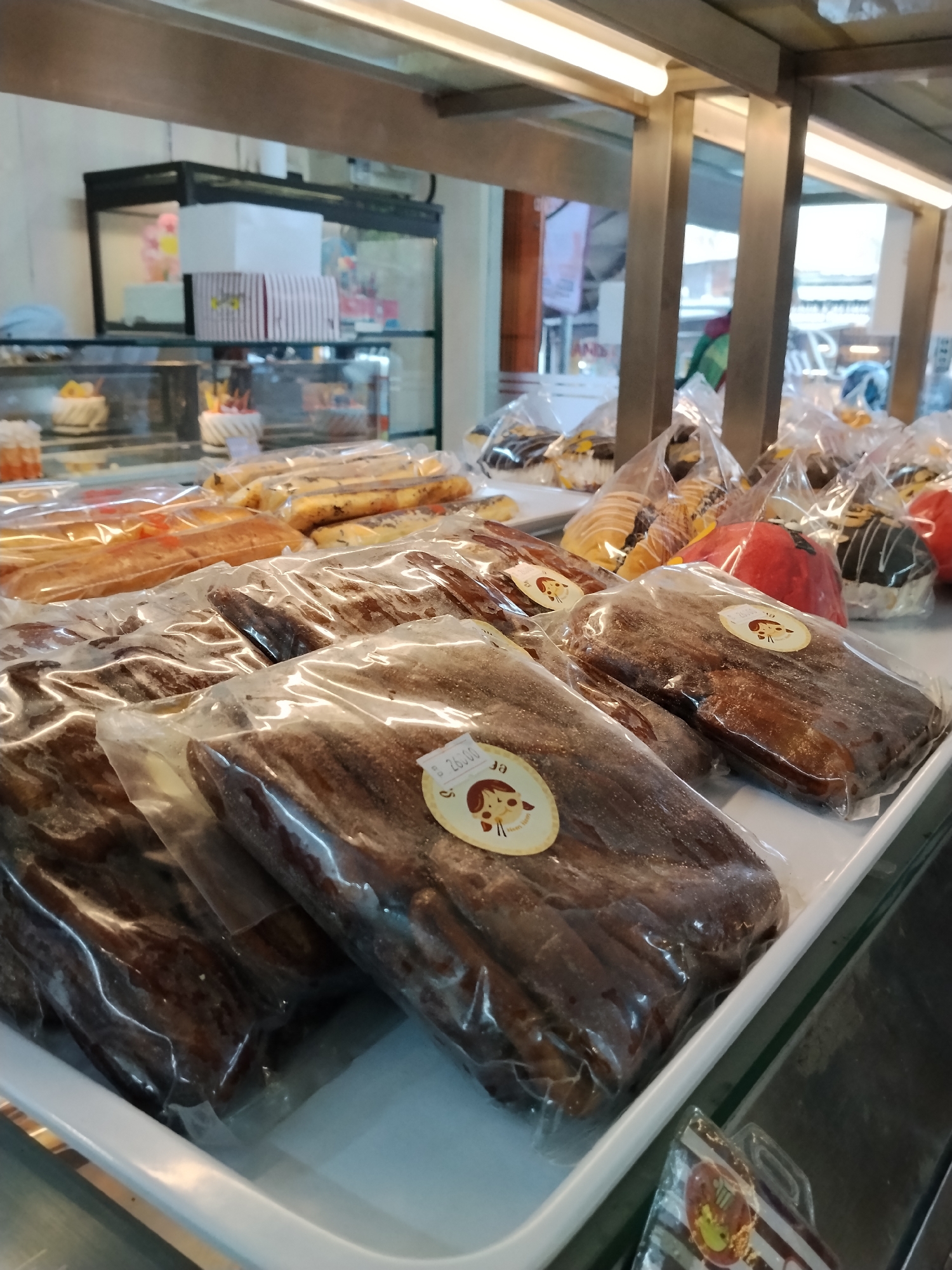 This is a close-up, eye-level shot of a display case filled with various baked goods, likely at a bakery or pastry shop in Ciruas, Indonesia. The foreground is dominated by several rows of individually wrapped, dark-colored breadsticks or pastries, some with a sticker featuring a cartoon dog. Behind these, further rows of similarly packaged baked goods, including croissants and what appear to be decorated cakes or donuts, are visible. The display is lit by bright overhead fluorescent lights, reflecting off the plastic packaging. In the background, shelves are visible with more boxes and what looks like a display of cakes, suggesting a wider variety of products offered. The overall impression is of a well-stocked and inviting bakery.