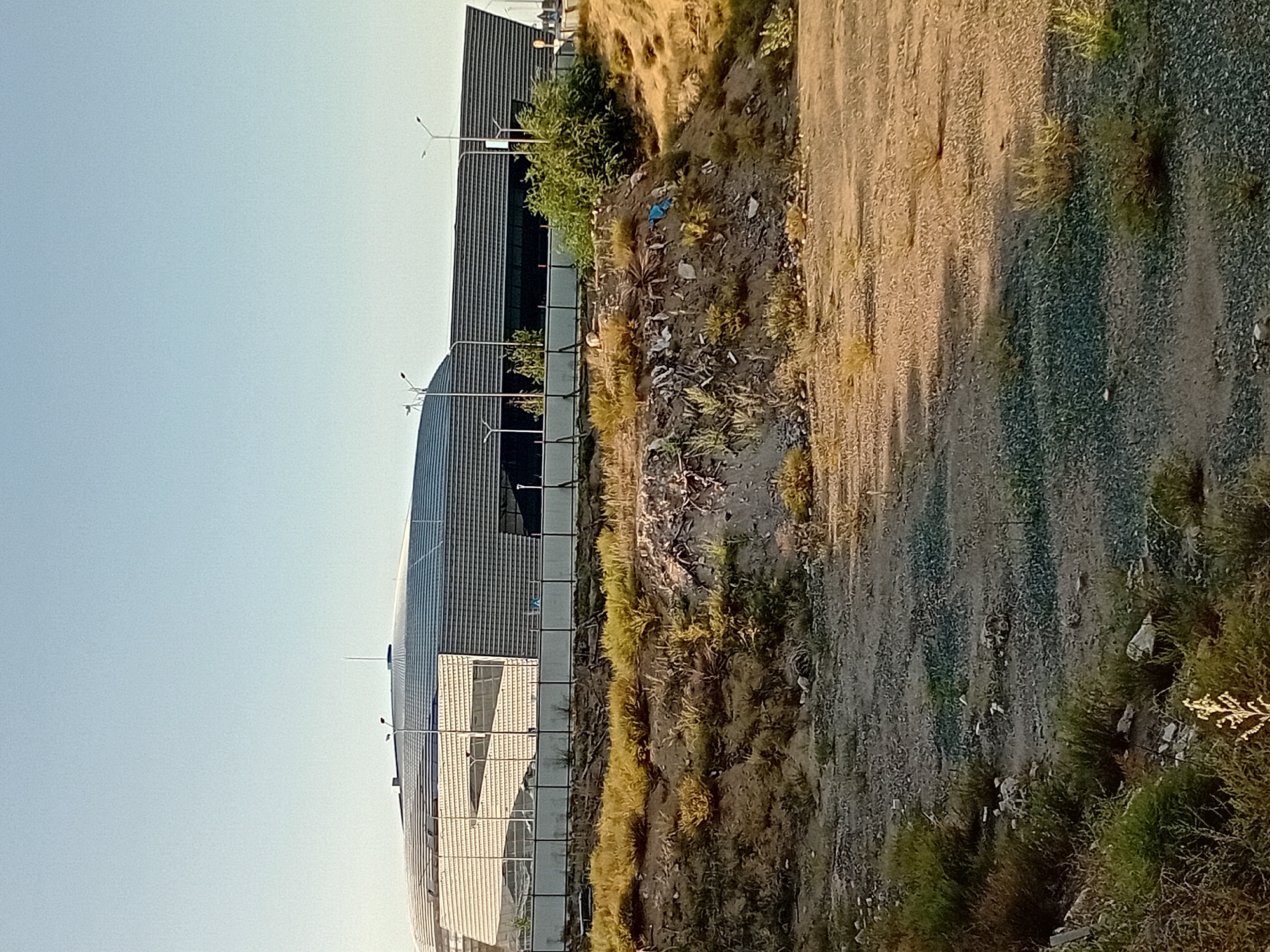 This is a daytime, eye-level, outdoor, full-shot photograph of the Semey Arena in Semey, Kazakhstan. The building is modern with a curved, metallic roof and a glass facade. It is situated behind a fence. In the foreground, there is a barren, uneven terrain with sparse vegetation and debris. The sky is clear and pale blue.