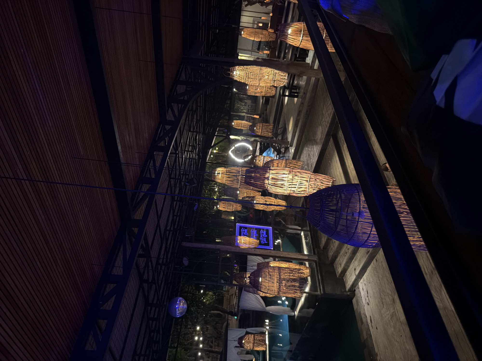 The image captures a low-angle perspective of a nighttime scene, likely in the Ubud District of Indonesia. The scene features a structure with a wooden slat ceiling and black metal framework. Hanging from the ceiling are numerous woven basket-like light fixtures of varying sizes and colors. A circular neon light can be seen in the distance. A blue neon sign with Asian characters is also visible. At the bottom of the image, a glimpse of trees and string lights suggests an outdoor setting. The image appears to be taken in a restaurant or bar with some patrons near the right-side frame, where blue lighting highlights some details of a person's clothing. The overall environment is dimly lit.
