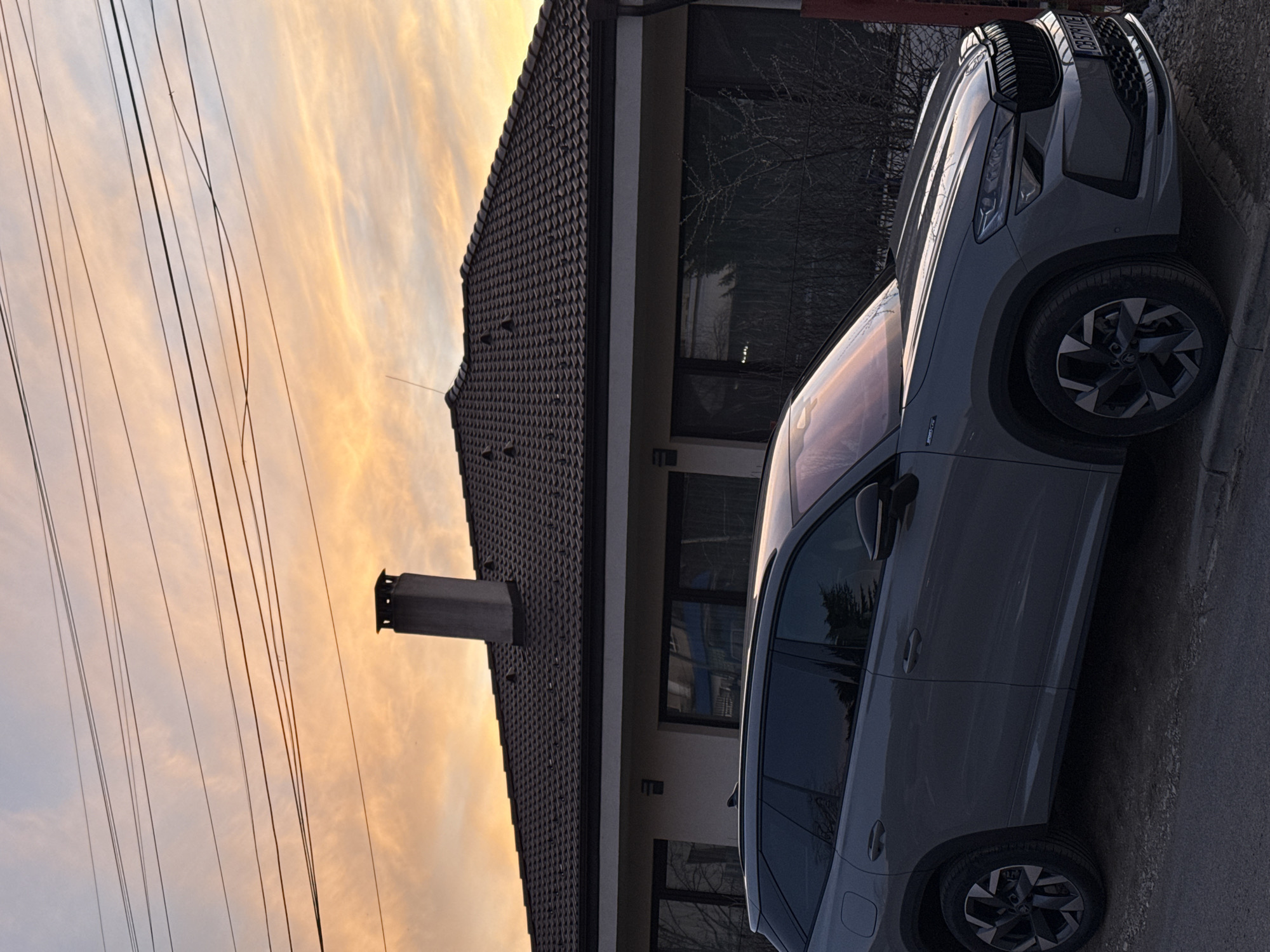 The image shows a grey Škoda Karoq SUV parked beside a house. The house has a dark brown tiled roof and a light-colored facade with windows. There is a chimney on the roof. Power lines run across the upper left of the image. The sky in the background is orange and yellow, suggesting either sunrise or sunset. Based on the license plate (CB 5701 CE), the car is registered in the Sofia region of Bulgaria. The location appears to be residential. The image lacks direct evidence that locates it in Etropole. If the car is located in Etropole, it's parked next to a residential building there. No people are visible in the photo.