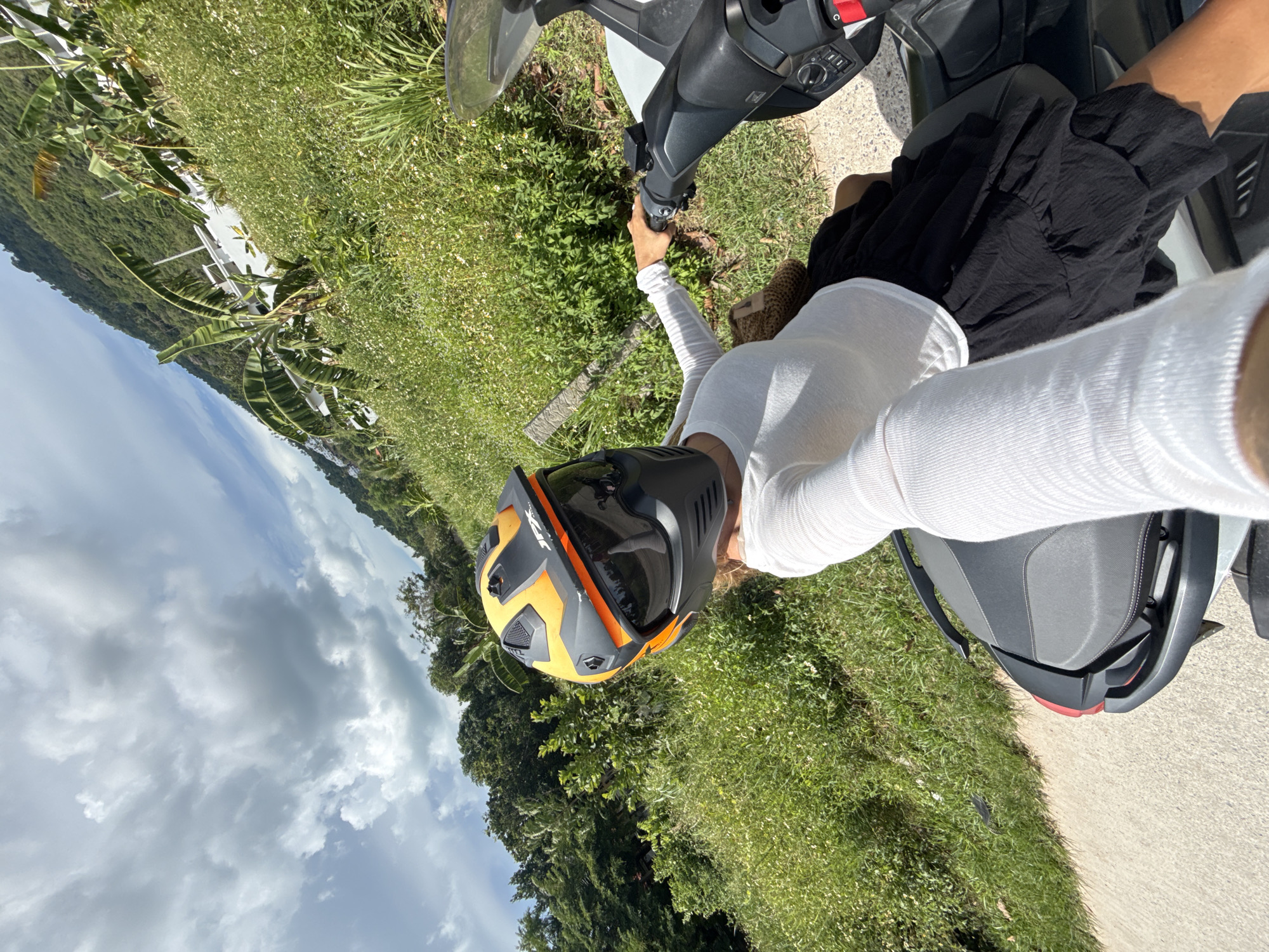 A person wearing a white long-sleeved shirt, a black skirt, and an orange and black helmet with a dark visor is seated on a scooter. The person is holding the handlebars with their right hand and appears to be taking a selfie. The scooter is on a paved road next to a grassy area with scattered white flowers and some taller green vegetation. In the background, a green, tree-covered hillside slopes upwards, with a white building visible partway up the slope. The sky is partly cloudy with white and gray clouds against a blue sky. The overall scene suggests a scenic outdoor location.