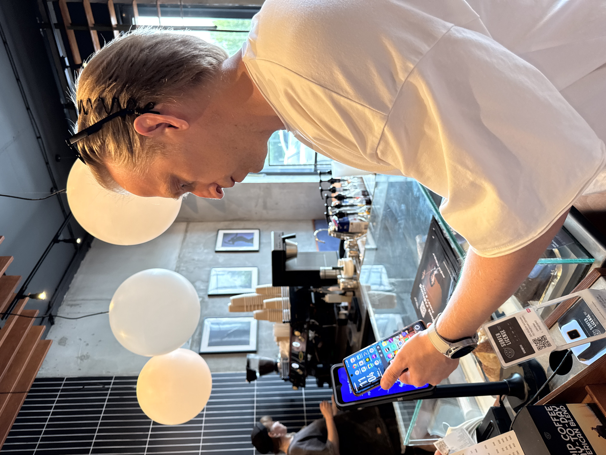 A person is looking at their phone in what appears to be a cafe in Tyumen, Russia, based on the text “Tyumen” in Russian on an item next to the phone. The person is wearing a white t-shirt and has a white smartwatch on their left wrist. They are holding a blue smartphone in their left hand and pointing at the screen with their right index finger. The phone screen displays various application icons. Next to the phone are objects with text related to coffee and surfer licenses, which includes QR codes. In the background, there is coffee-making equipment, cups, and pictures on the wall. In the lower part of the scene, a person wearing a cap is partly visible. The setting includes white balloon-like light fixtures hanging from the ceiling and a tiled black-and-white floor.