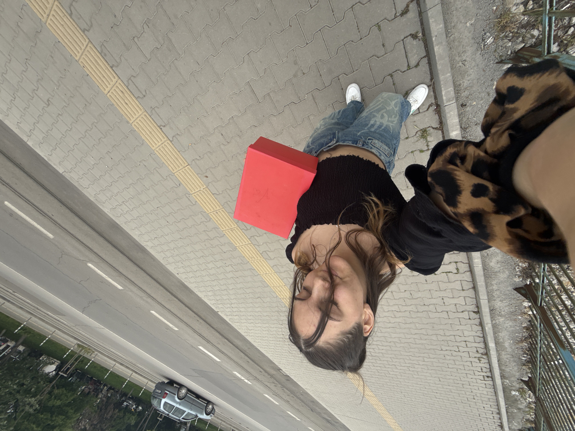 A woman stands on a sidewalk adjacent to a multi-lane road, possibly a highway, as a car travels in the left lane, as viewed from the perspective of the image. Tactile paving is visible on the sidewalk to the left. The woman is holding a red box. She wears white sneakers and faded blue jeans. She is also wearing a black top. A garment with an animal print pattern is next to her arm. A metal fence runs along the edge of the sidewalk. Based on general characteristics and street design, the location appears to be in or around the city of Sofia, Bulgaria.