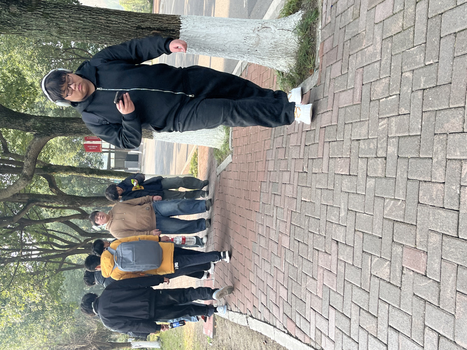 A group of students walk on a brick pathway lined with trees. A person in a black tracksuit and headphones stands on the right, looking at a phone. Tree trunks are painted white at the base. The pathway is paved with alternating brick patterns. Some students wear backpacks. Some of the trees are leafless.